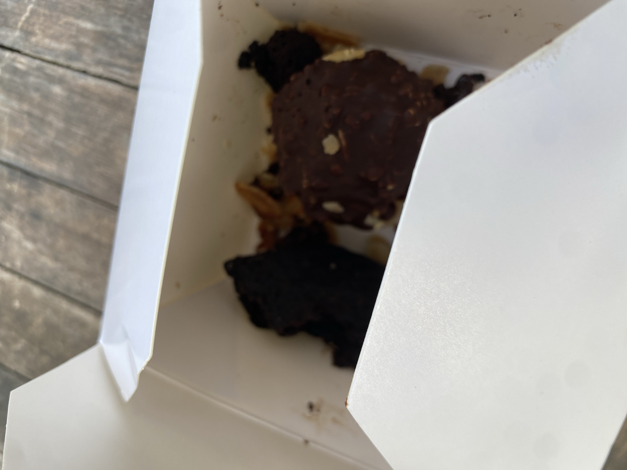 Two pieces of dessert are in a white cardboard box. One piece is a dark brown brownie. The other piece is a chocolate-covered item with nuts and a light-colored filling. The box is open and sits on a wooden surface.
