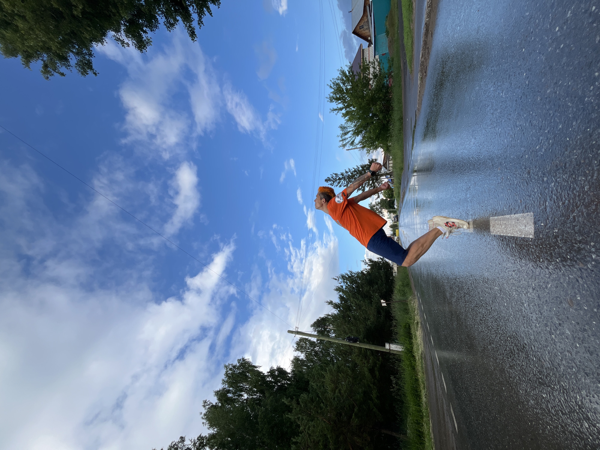 A young man with orange hair is captured mid-air, performing a backbend on a wet asphalt road. He is wearing an orange t-shirt and navy blue shorts, with white sneakers. The road, marked with a white stripe, reflects the bright blue sky filled with scattered white clouds. Lush green trees line the sides of the road, and houses with visible roofs are present in the background, suggesting a residential area. A utility pole with wires stretches across the frame, contributing to the rural or suburban setting. The overall scene is dynamic, with the man's pose suggesting an athletic or acrobatic movement. This scene is located in the city of Chaikovsky, Russia.