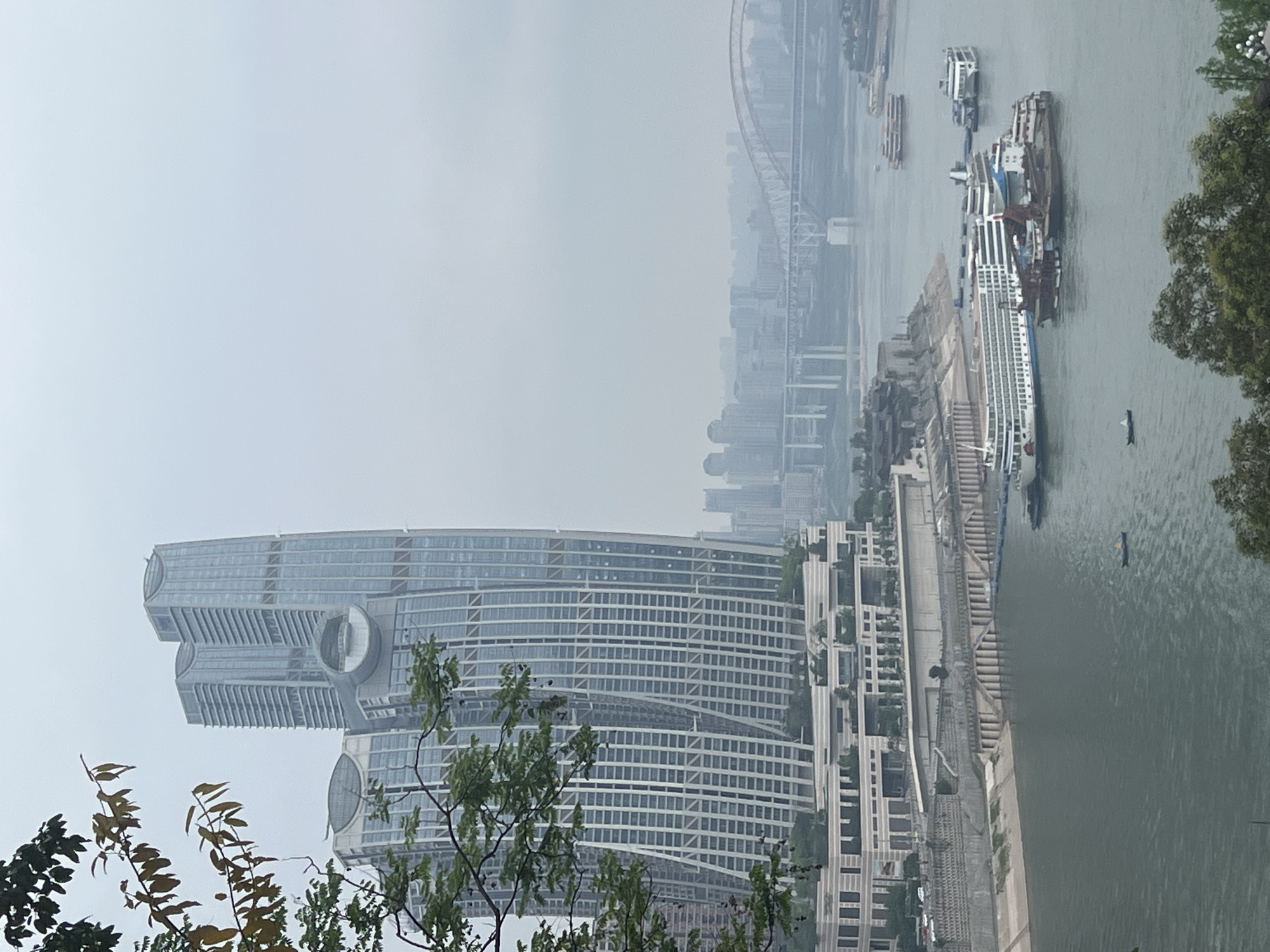 This photograph depicts a cityscape in Nan'an District, China, under hazy skies. Prominently featured is a modern, high-rise building with a distinctive design, including circular architectural elements. A river flows alongside the city, with boats and barges visible on the water. A bridge can be seen in the distance, obscured by the haze. Trees and foliage are present in the foreground, framing the urban landscape.