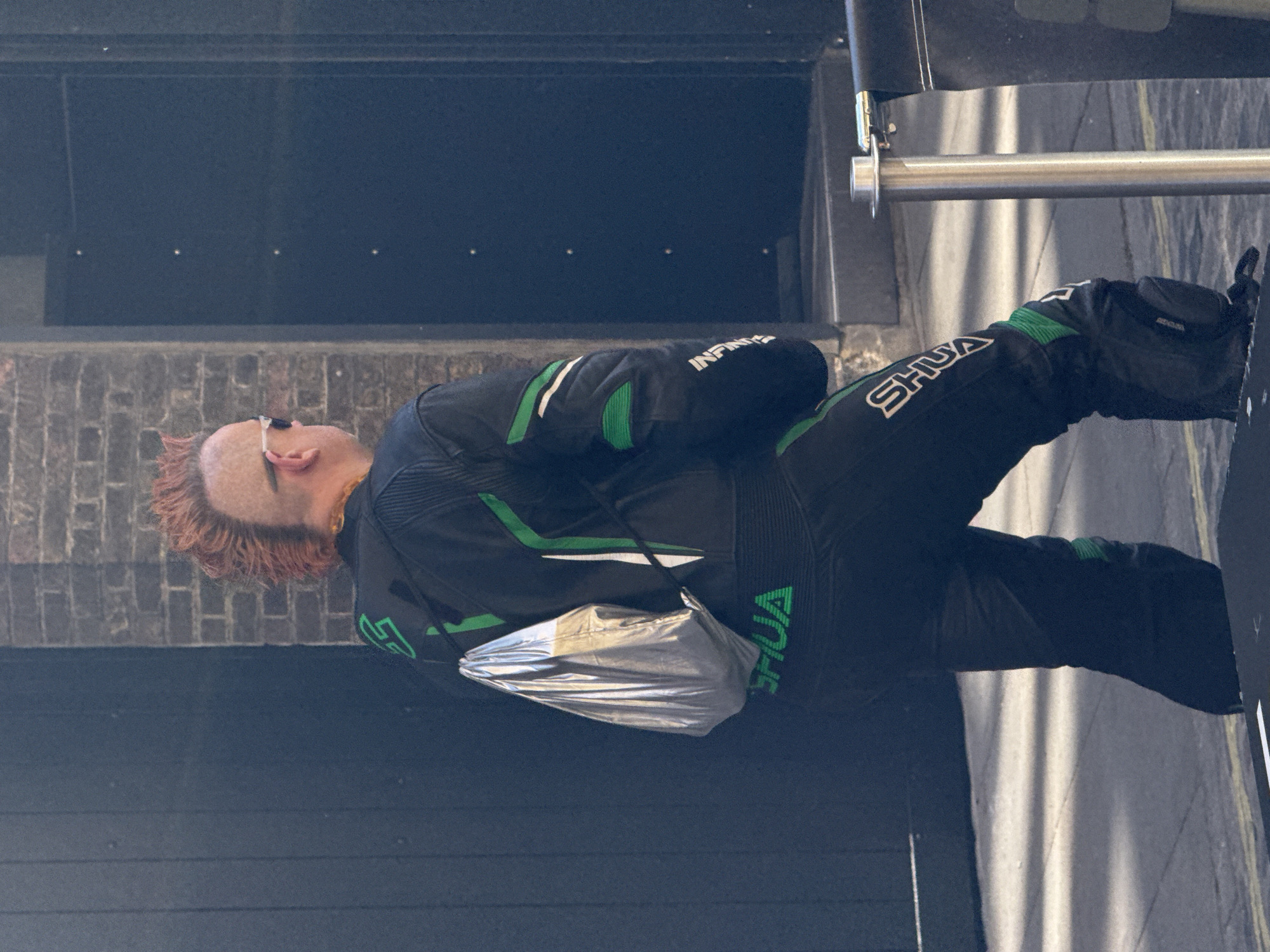The image features an adult individual in an urban setting, possibly in London, United Kingdom, captured in bright daylight. The person has reddish-orange hair and wears sunglasses pushed up onto their head. They are dressed in a black, form-fitting motorcycle racing suit, accented with striking green stripes and panels. The suit prominently displays the brand name "INFINITI" in white lettering on the right arm, and "SHUA" on the left arm and left leg. A reflective, silver-colored pouch or bag hangs from their waist area.
The individual is standing or leaning, with their head tilted upwards and slightly to the right. The background on the left consists of a dark brick wall with a darker, smooth surface above it. On the right side of the frame, a metal railing or bar is visible. The ground is dark asphalt with subtle light-colored markings and strong shadows, indicating sunny weather. No specific activity is taking place beyond the person's pose.