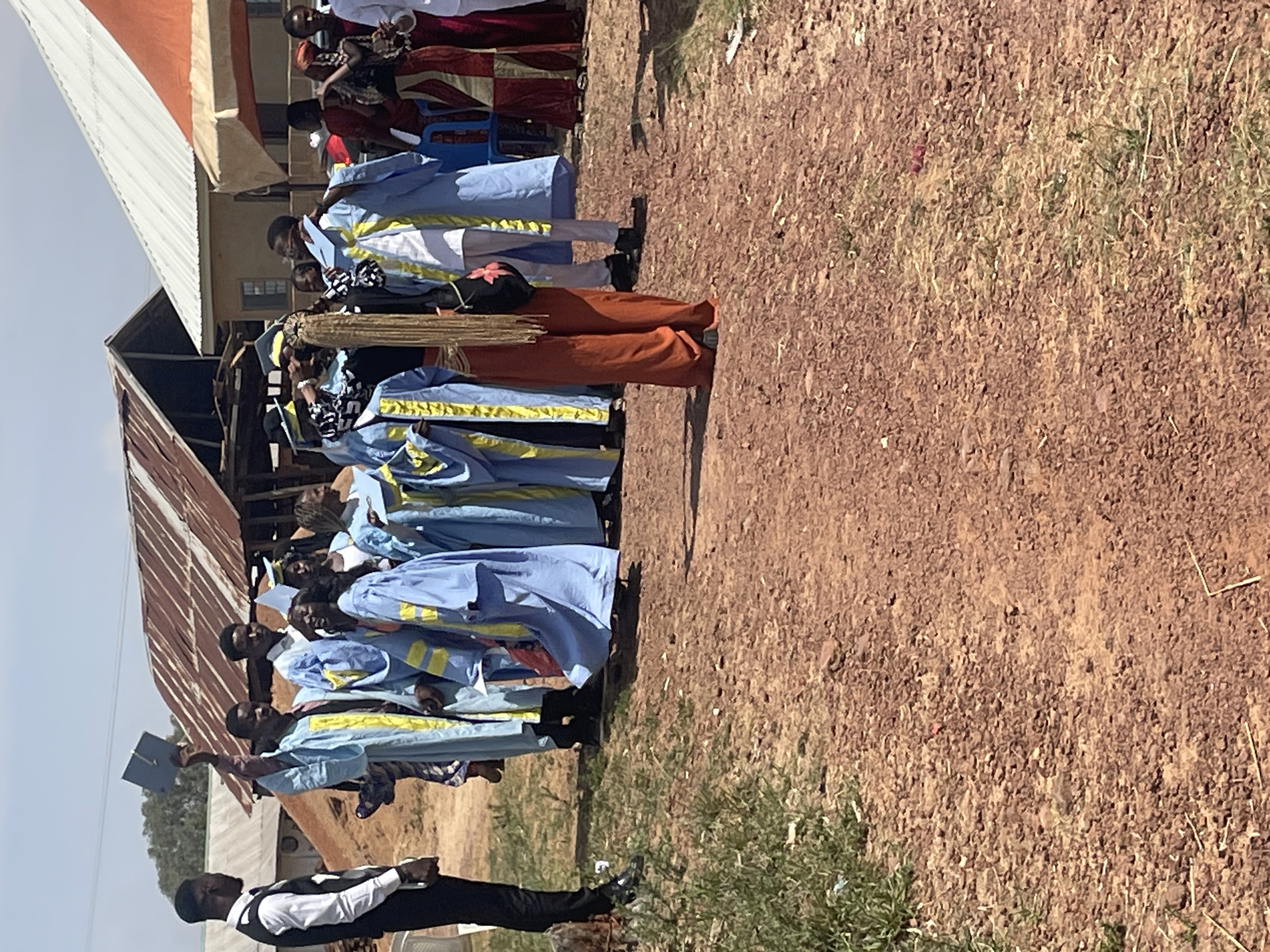 The image depicts an outdoor graduation ceremony taking place in Durimi, Nigeria. A group of graduates is wearing blue and yellow gowns. Some individuals are holding up graduation caps. A man dressed in a white vest and black pants is standing facing them. The setting includes a building with a corrugated metal roof and reddish-brown dirt ground. A small amount of vegetation is visible on the ground.