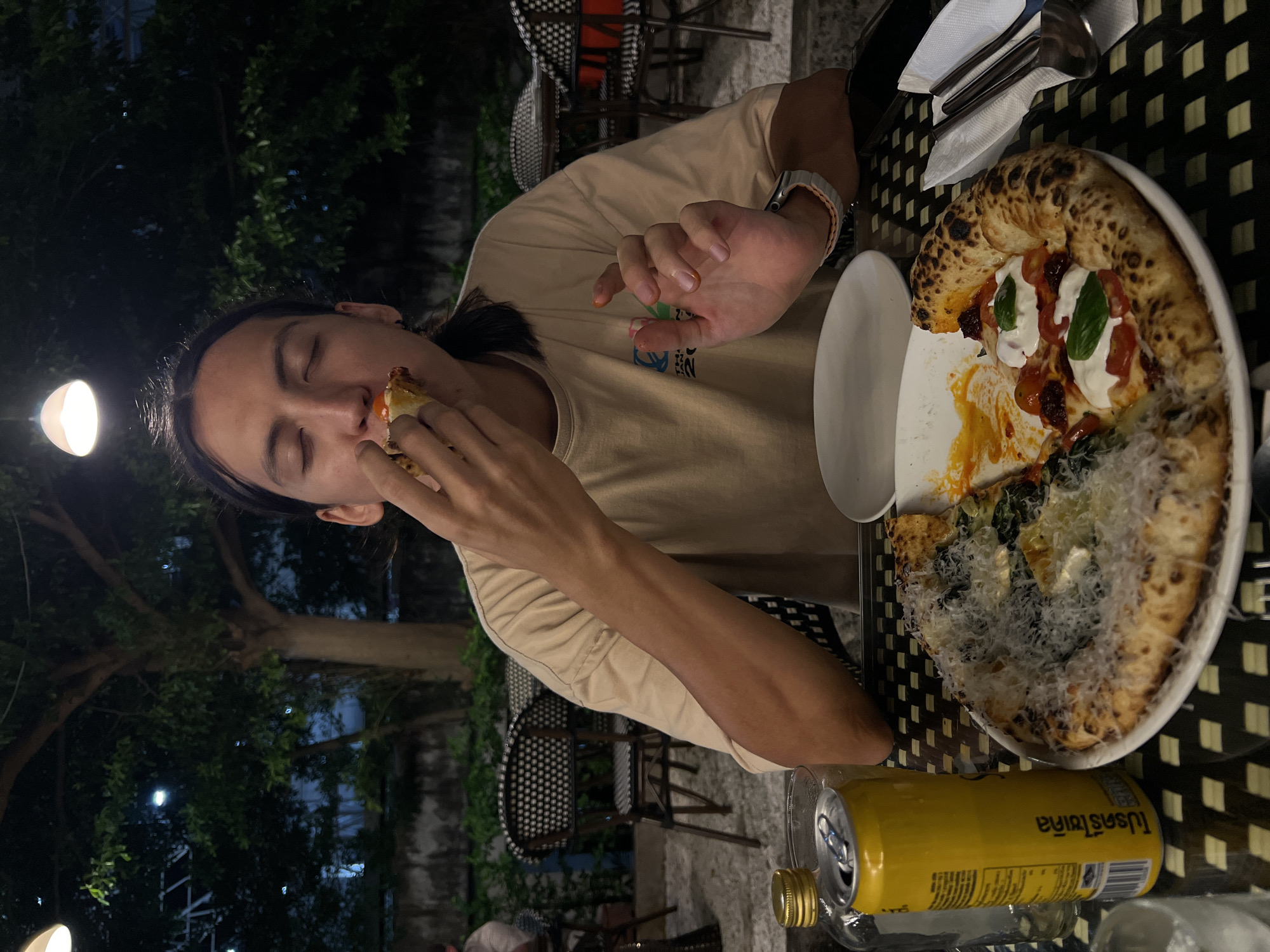A person is eating a slice of pizza at an outdoor table in Unknown, Unknown. The person has long dark hair and is wearing a beige shirt and a watch. They are holding a slice of pizza to their mouth with their right hand. On the table is a plate with a pizza cut in half. One half has tomato sauce, mozzarella, and basil, and the other half has spinach and cheese. There is also a small white plate, silverware, and a yellow can with Thai writing on it. The table has a black and white checkered pattern. In the background, there are trees, chairs, and a building.