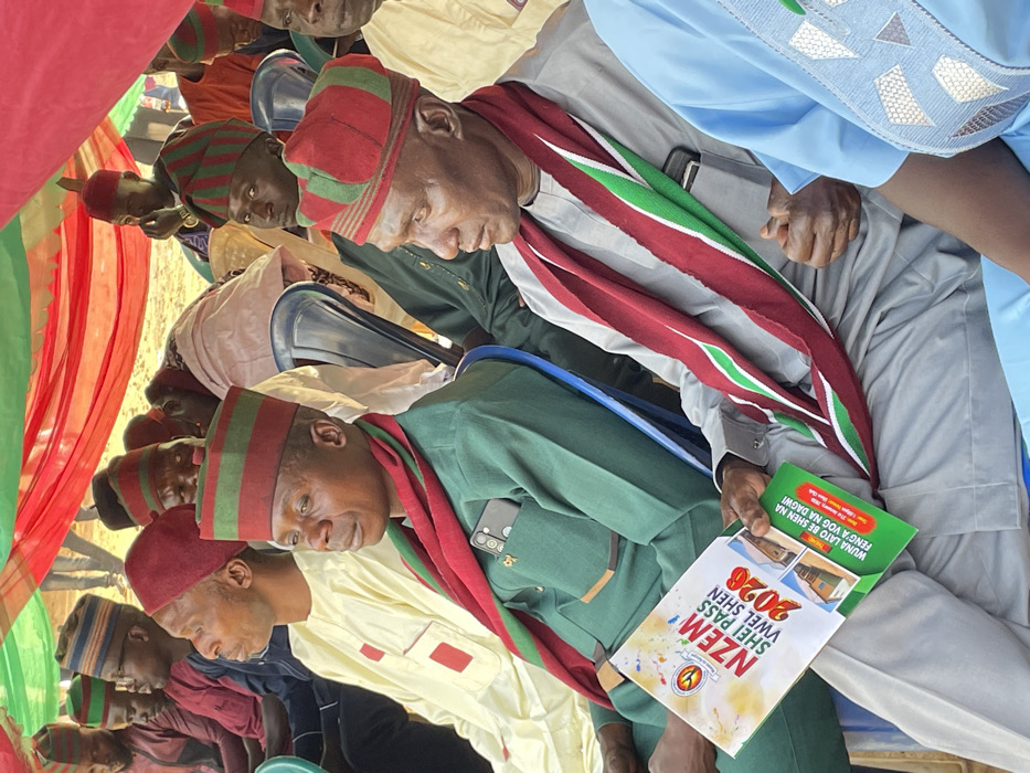 The image captures a gathering of several men, predominantly of African descent, seated and attending an event, likely indoors or under a tent, given the draped fabrics visible in the background. The photograph is oriented vertically, with subjects appearing to be leaning to the right.
The men are mostly dressed in matching traditional attire, featuring distinctive red and green striped caps and corresponding scarves or stoles draped over their shoulders. Some wear green military-style uniforms underneath these traditional accessories, while others are in light-colored robes, such as a pale yellow kaftan and a light blue robe with intricate embroidery on the chest and sleeve.
In the foreground, a man wearing a green uniform, a red and green striped cap, and a matching scarf is holding a white pamphlet or booklet in his left hand. A mobile phone with multiple camera lenses is tucked into the breast pocket of his uniform. Another man seated next to him, wearing a light grey robe and a similar cap and scarf, is also visible. His right hand is holding what appears to be another green pamphlet.
The pamphlet held by the man in the foreground has prominent text and graphics. Large text at the top reads "NZEM," followed by "SHEI PASS," "VWEL SHEN," and a large "2026." Below this, there's a circular logo featuring a stylized white symbol against a darker background, possibly a 'Y' or similar shape. Further down, there are smaller images of what appear to be houses or buildings. The green pamphlet held by the other man contains text that reads "WUNA LATO BE SHEN NA FENG'A VOG NA DAGWI." The overall design suggests promotional material for an upcoming event or campaign slated for 2026, possibly related to community development, elections, or a cultural festival, given the traditional attire.
The background shows red and green striped fabric draped vertically, consistent with a decorated event space or a large tent. The overall atmosphere appears to be formal, suggesting a meeting, ceremony, or political rally. The men's expressions are generally attentive, with some looking down at their pamphlets and others looking forward. The lighting suggests it is daytime.