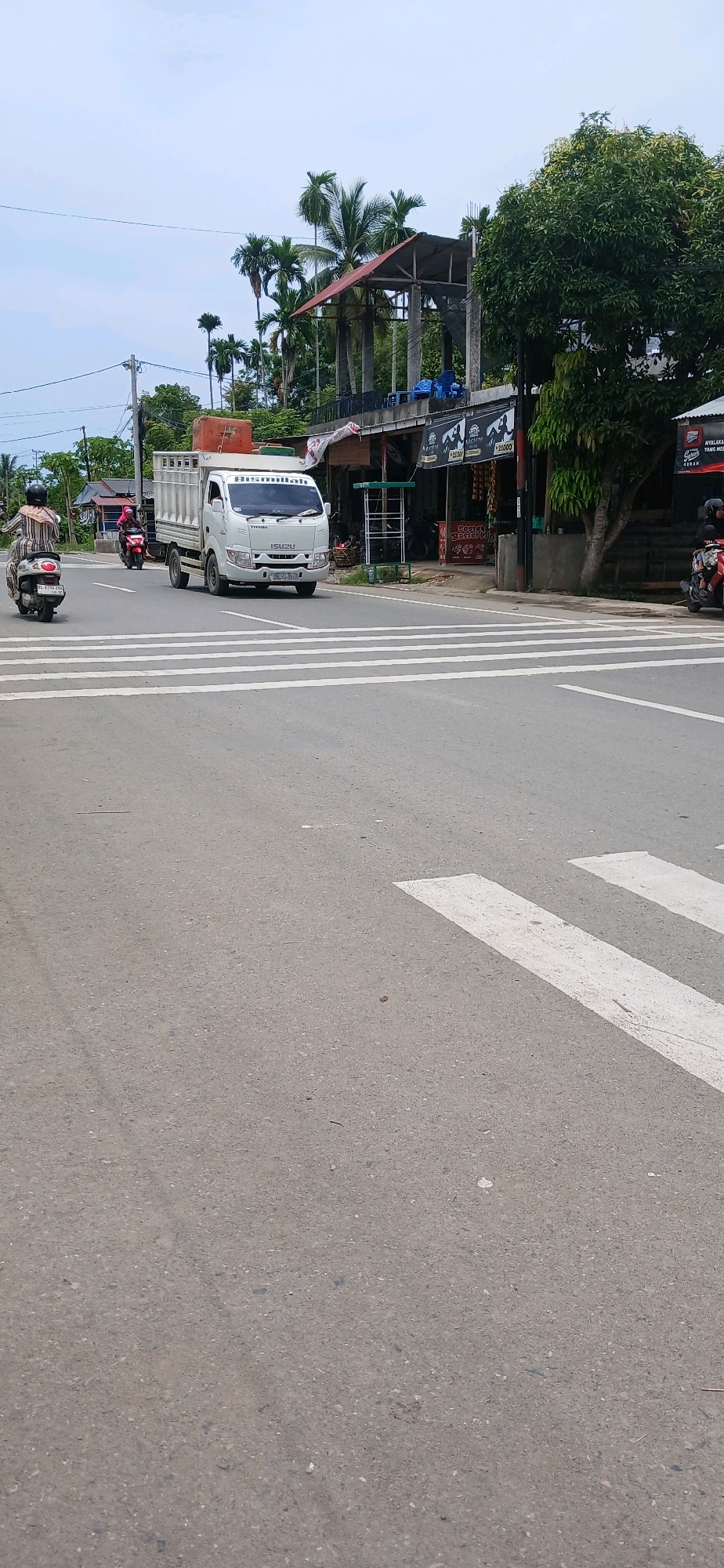 In Matangjareung, Indonesia, a white Isuzu truck with an orange container on its bed is crossing a pedestrian crossing on a paved road. Two individuals on motorcycles are also present: one on a scooter to the left of the truck, and another on a motorcycle to the right of the truck, further down the road. In the background, there are several buildings, including a structure with a red roof and concrete pillars, and other storefronts with signage. Palm trees and other lush greenery are visible, indicating a tropical or subtropical climate. The sky is overcast with light clouds.