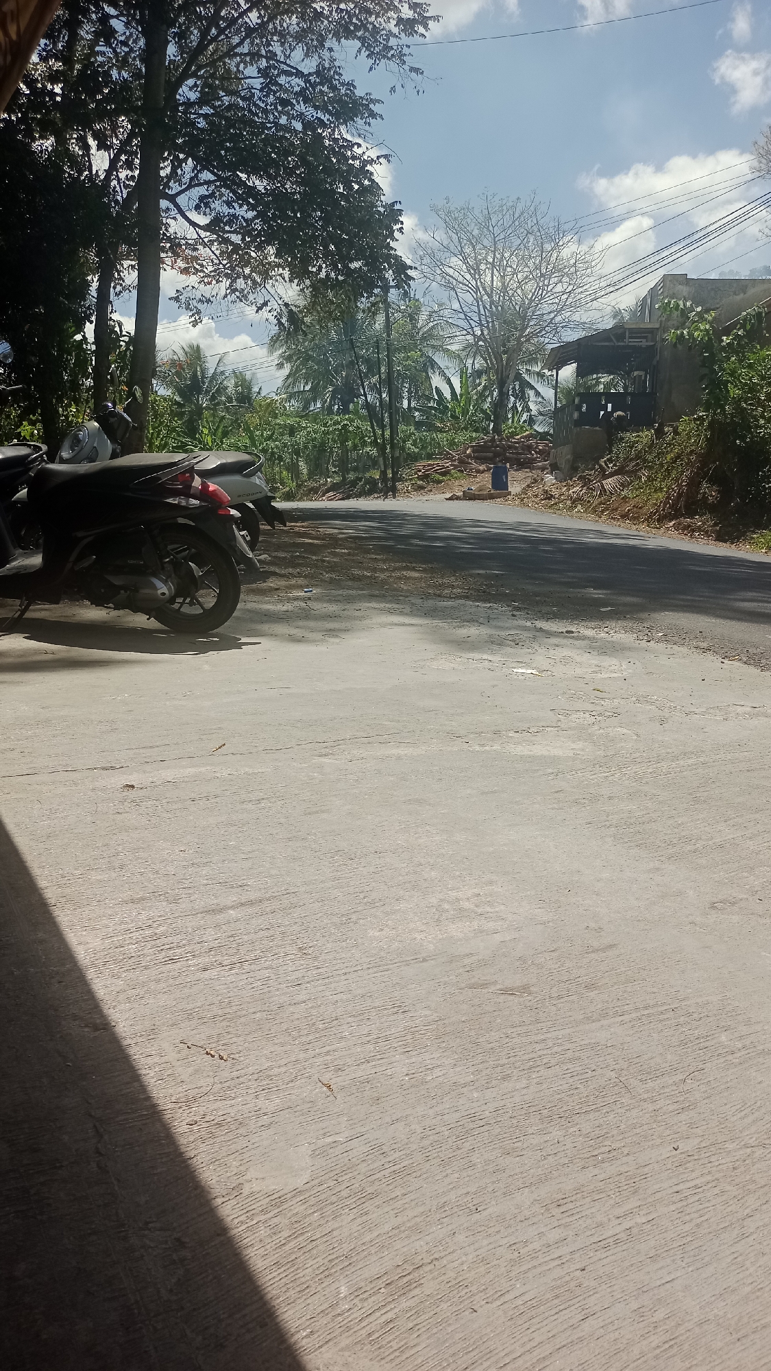 A motorcycle is parked on a concrete surface in front of a paved road. Trees and vegetation are visible in the background, along with a small building and power lines. The sky is blue with scattered clouds. The scene appears to be in a rural or semi-rural area, possibly in Cikalong, Indonesia, given the tropical foliage.