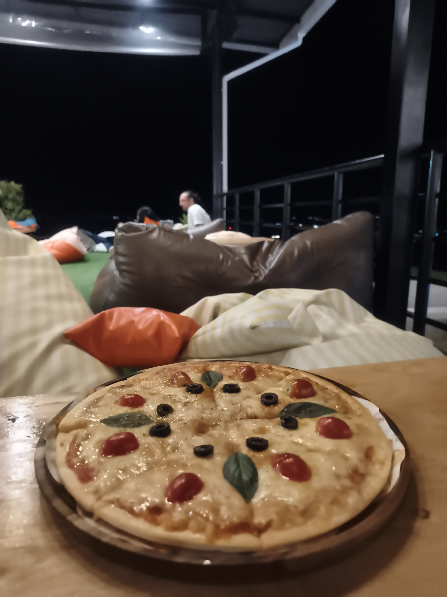 A pizza sits on a wooden table in the foreground, topped with cherry tomatoes, black olives, and basil leaves. The pizza is round with a golden crust and melted cheese. In the background, beanbag chairs are scattered on a grassy area under a covered patio. Several people are visible lounging on the beanbags. The scene appears to be outdoors at night, suggested by the dark sky and the lighting from the patio. The overall atmosphere seems relaxed and casual. The location is identified as Baan Bang Rak, Thailand.