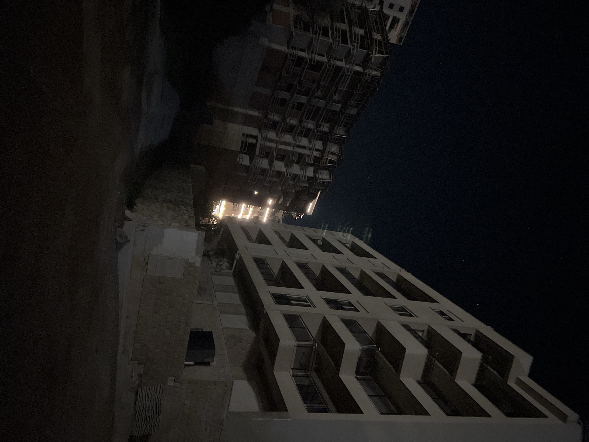 The image shows two buildings against a dark night sky, potentially located in Boreti, Montenegro. One building features a facade with balconies in a zigzag pattern. The other building is a taller, modern structure. Both are illuminated by internal and external lights. The environment appears urban, with the buildings dominating the frame.