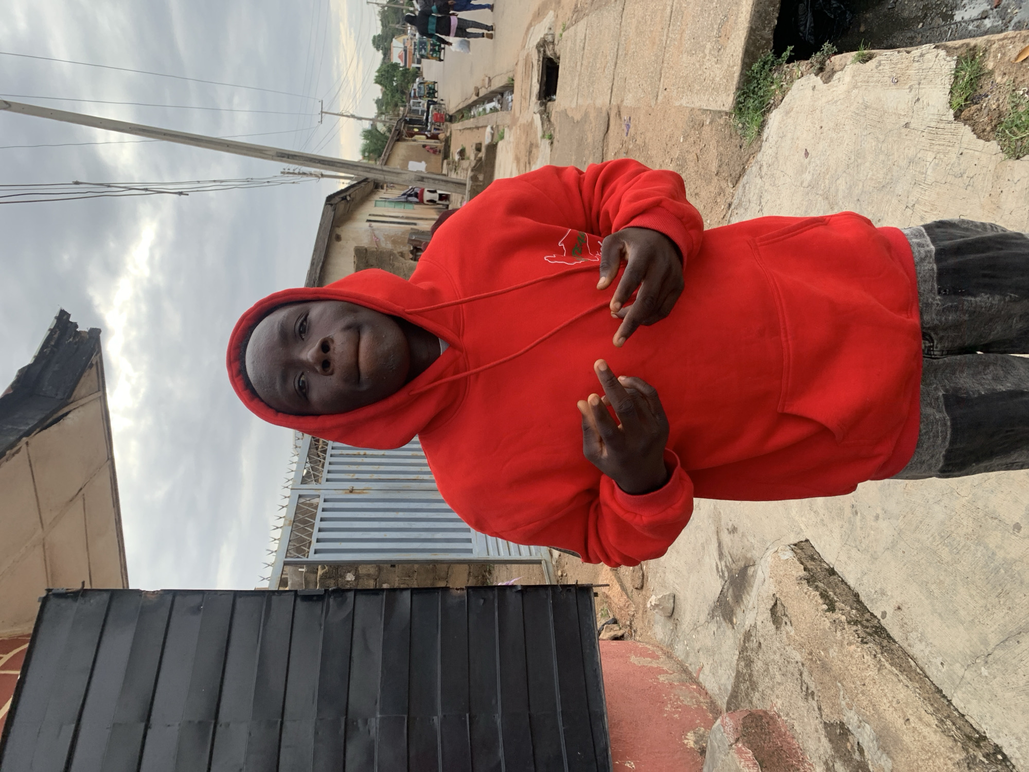 This image captures a young male subject standing on a street in the city of Jos, Nigeria. The subject, positioned slightly to the left of the frame's center, is a person with dark skin, wearing a bright red hooded sweatshirt with the hood pulled up. His hands are raised in front of him, each forming a V-sign with the index and middle fingers, palms facing forward. His facial expression is neutral, with slightly pursed lips. He appears to be wearing dark, possibly acid-washed or distressed, jeans.
To the immediate left of the subject is a dark, horizontally-ridged metal structure, partially obscuring a light-colored building with a gray metal gate topped with barbed wire in the background. A red-painted concrete structure is visible at the bottom left of the frame. The foreground consists of a cracked and uneven concrete sidewalk, bordered by an open drainage ditch containing dark water and debris.
The street extends into the background, lined with various buildings of mixed construction and finish. Utility poles with multiple overhead wires are prominent along the street. Further down the road, several vehicles, including tuk-tuks, and pedestrians are visible. The sky is uniformly overcast with gray clouds.