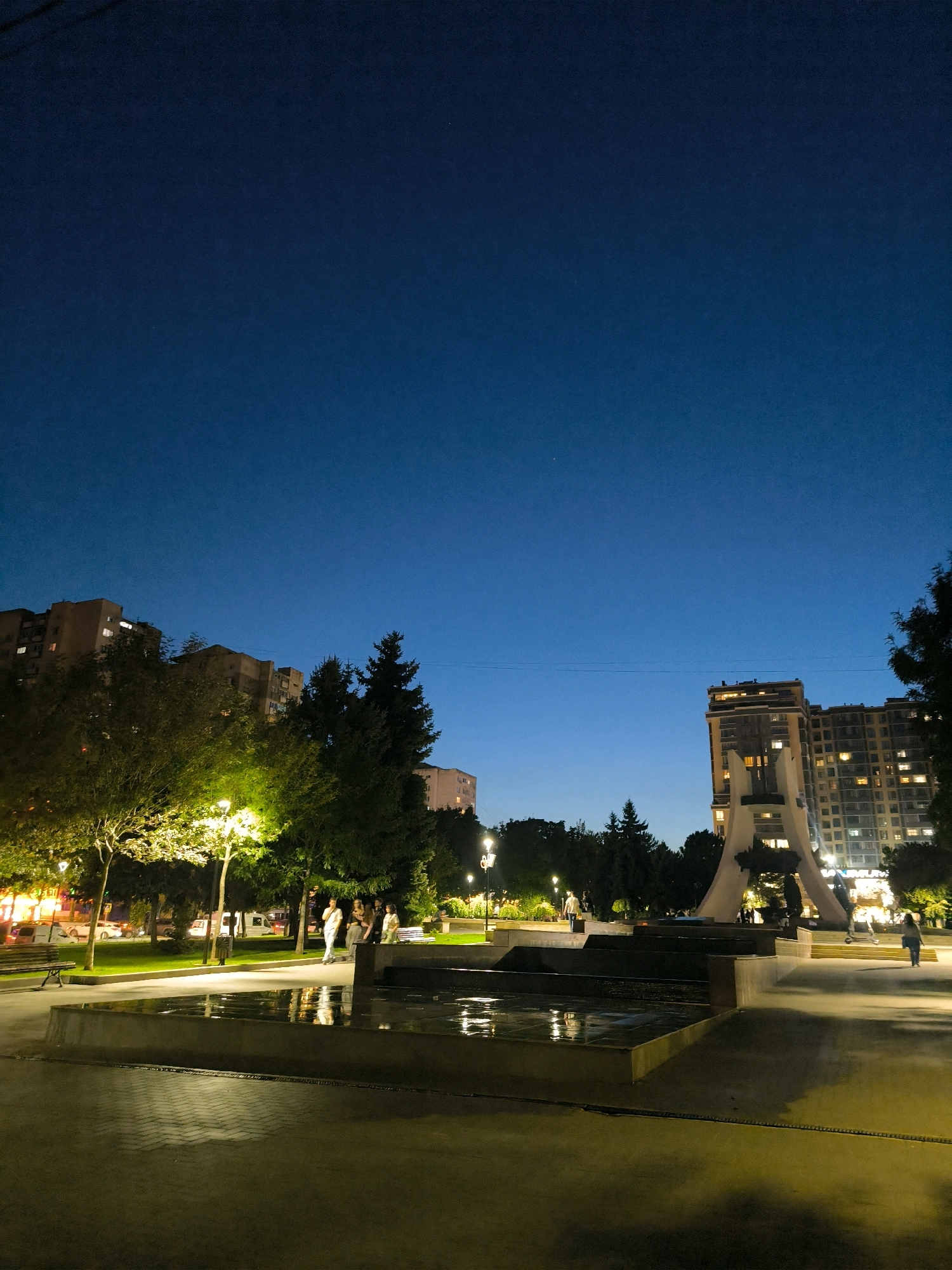 The image depicts a nighttime scene in a public square in Chișinău, Moldova. The sky is a deep twilight blue, transitioning to black at the top of the frame. A prominent modern sculpture, resembling a large, abstract white structure with orange lighting, stands to the right, flanked by tall residential buildings with illuminated windows. In the foreground, a multi-tiered water fountain, filled with dark water reflecting the surrounding lights, sits in the center of a paved plaza. To the left, trees with green foliage are illuminated by streetlights, casting a warm glow on the surrounding park area. Several individuals are visible: a group of people stand near the fountain on the left, appearing to be engaged in conversation, while a lone figure walks away from the camera towards the right. Another person is seen walking in the background near the sculpture. In the distance, vehicles can be seen on a street bordering the park. The overall atmosphere is tranquil and urban, suggesting a public space that is active even after dark.