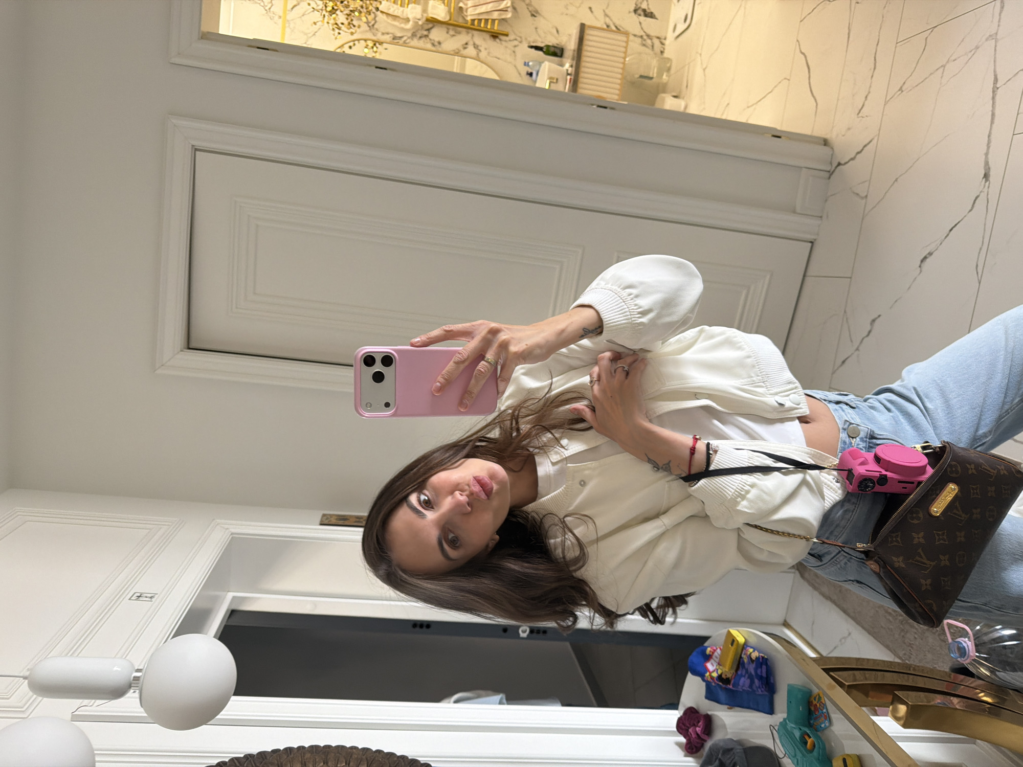 A woman is taking a selfie in a bathroom. She is wearing a white cardigan, blue jeans, and carrying a brown Louis Vuitton bag with a pink camera attached. The bathroom has white marble walls and a white door. The woman is looking at the camera with a pout. The location cannot be determined to be Krasnoyarsk, Russia from the image.