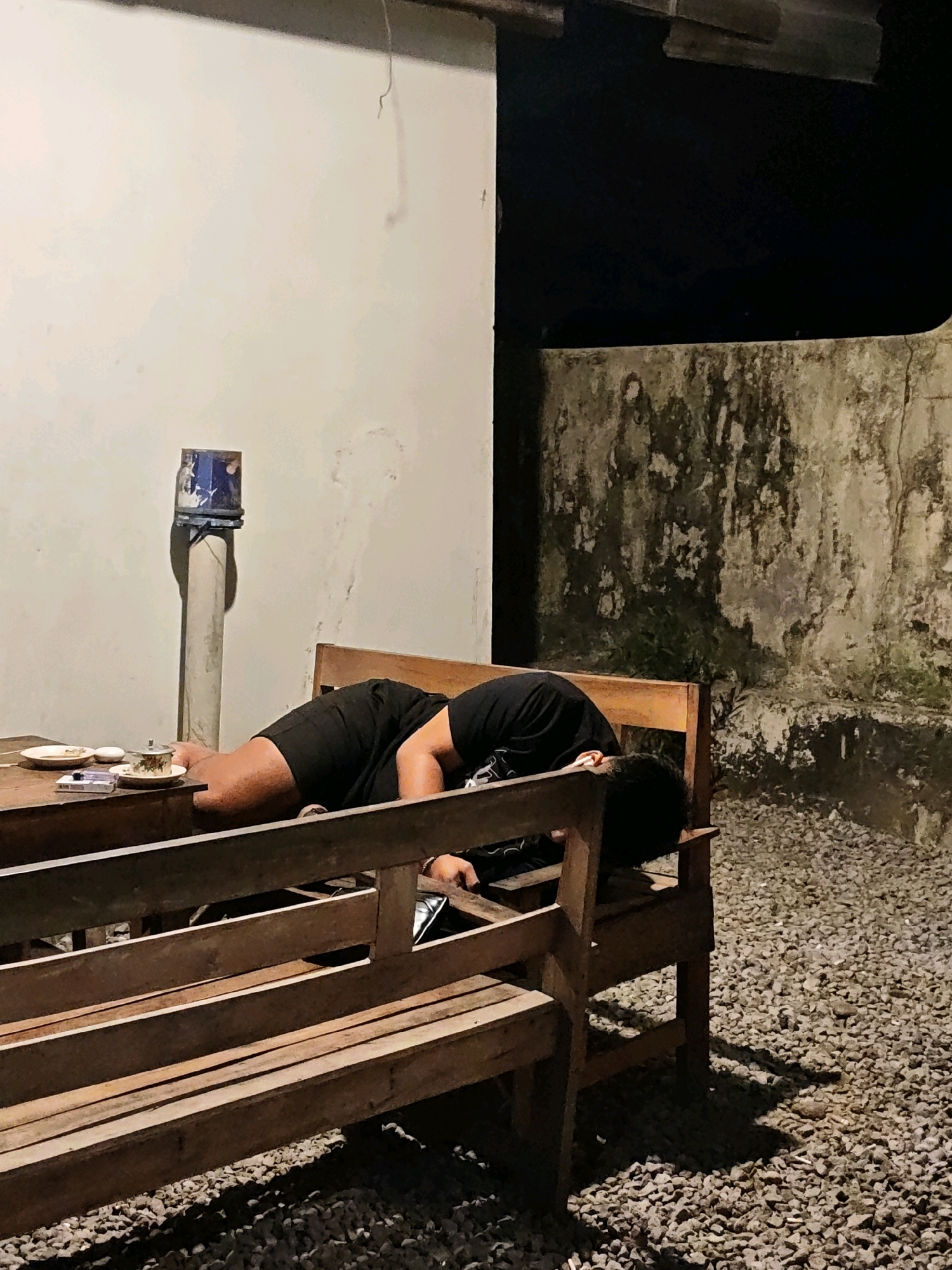 A person lies asleep face down on a wooden bench at an outdoor table. The person is wearing black shorts and a black t-shirt. A table in front of them holds a plate, a cigarette pack, and a small cup with a lid. A textured wall is behind the table, and the ground is covered in gravel. A pipe with a blue and white object attached to the top is also visible against the wall. The setting appears to be in Ngasem, Indonesia, likely a cafe or restaurant given the table and seating arrangement. The lighting suggests it is evening or night.