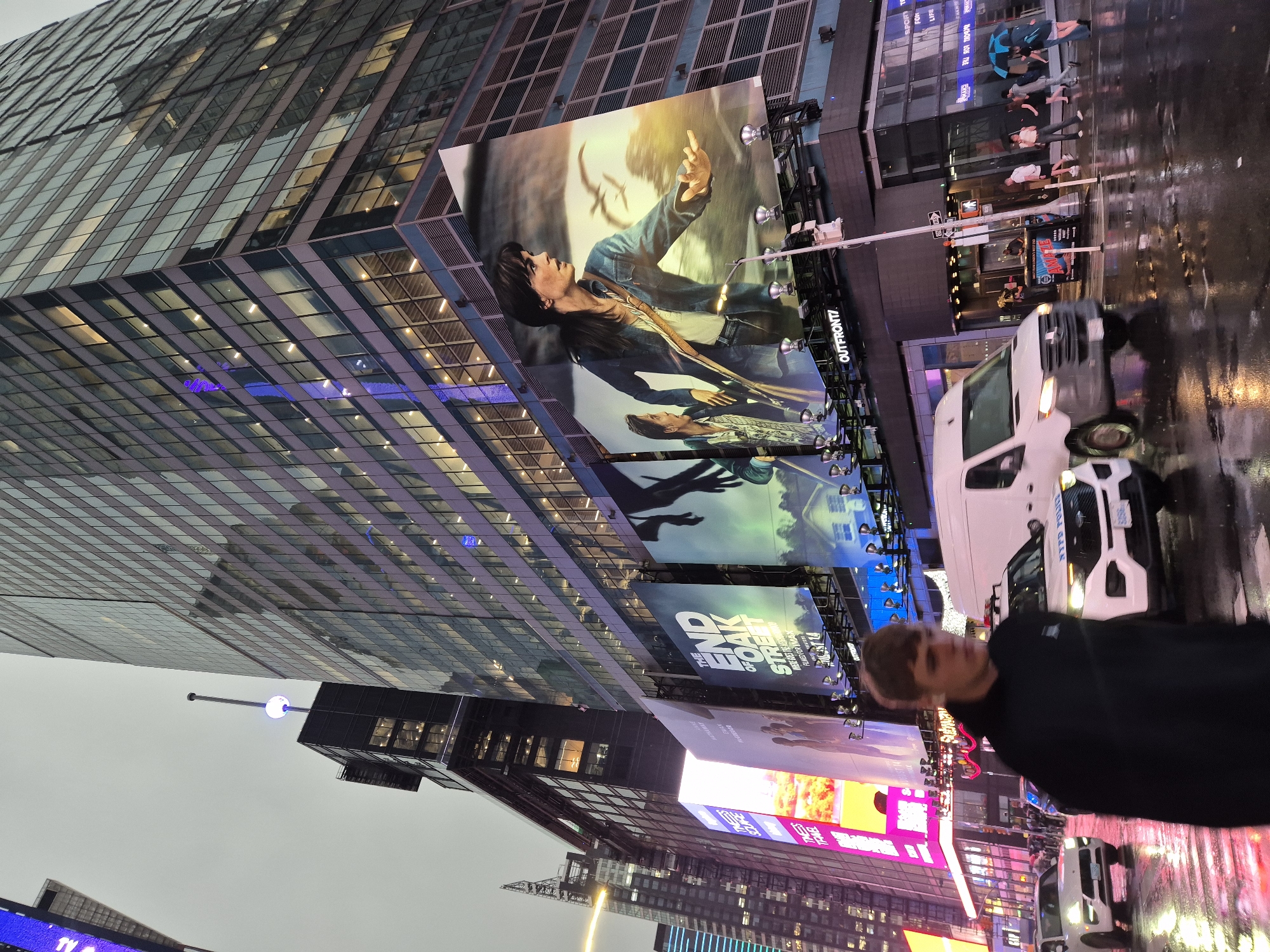 This image depicts Times Square in New York City, United States of America, on a cloudy and rainy day. The foreground features a young man wearing a black hoodie, walking towards the camera with his head down. He is partially obscured by a white van and a police vehicle with flashing lights. The street is wet and reflective, indicating recent rainfall.
In the background, a large, modern skyscraper dominates the scene, with its glass facade reflecting the surrounding buildings and the overcast sky. Several large digital billboards are mounted on the building, displaying advertisements. One prominent billboard features a movie poster with two actors. Other smaller billboards and signs are also visible.
Several other vehicles, including a white SUV and a white van, are on the wet street. Pedestrians, some holding umbrellas, are visible crossing the street and walking on the sidewalk. The overall atmosphere is urban and busy, characteristic of Times Square.