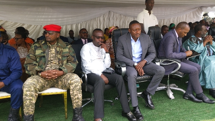 The image shows a group of men seated under a white canopy tent. The men are sitting on black, metal-framed chairs with black leather seats. The chairs are positioned on a green, artificial grass surface. The men are wearing various attire, including military fatigues, suits, and traditional clothing. The man in the centre is wearing a gray suit and black shoes, his hands clasped together in his lap. He is looking at the camera. The man to his right is wearing a blue suit, black shoes, and is looking down at a piece of paper he is holding. The man to his left is wearing a white shirt and black pants. He has his right hand near his mouth and is looking to his left. The man furthest to the left of the frame is wearing a camouflage jacket, red beret, and black pants. He is seated on a chair with a gold frame. He has both hands clasped in his lap. The man in the background is standing, wearing a white t-shirt. The setting is an outdoor event or ceremony in Rukoko, Democratic Republic of the Congo, at the location specified by the given coordinates.