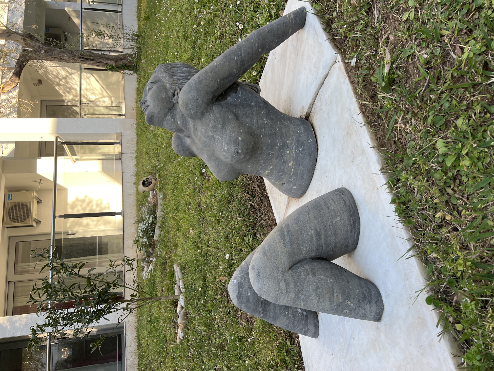 A partly-buried, grey, stone sculpture of a woman lies on white marble or stone slabs set into the grass. The sculpture is split into two pieces, a torso with one arm raised, and two legs bent at the knees. The location appears to be outdoors, near a building with large glass windows and doors. The ground is covered in grass and small white flowers. The image was taken in Brežine, Montenegro.