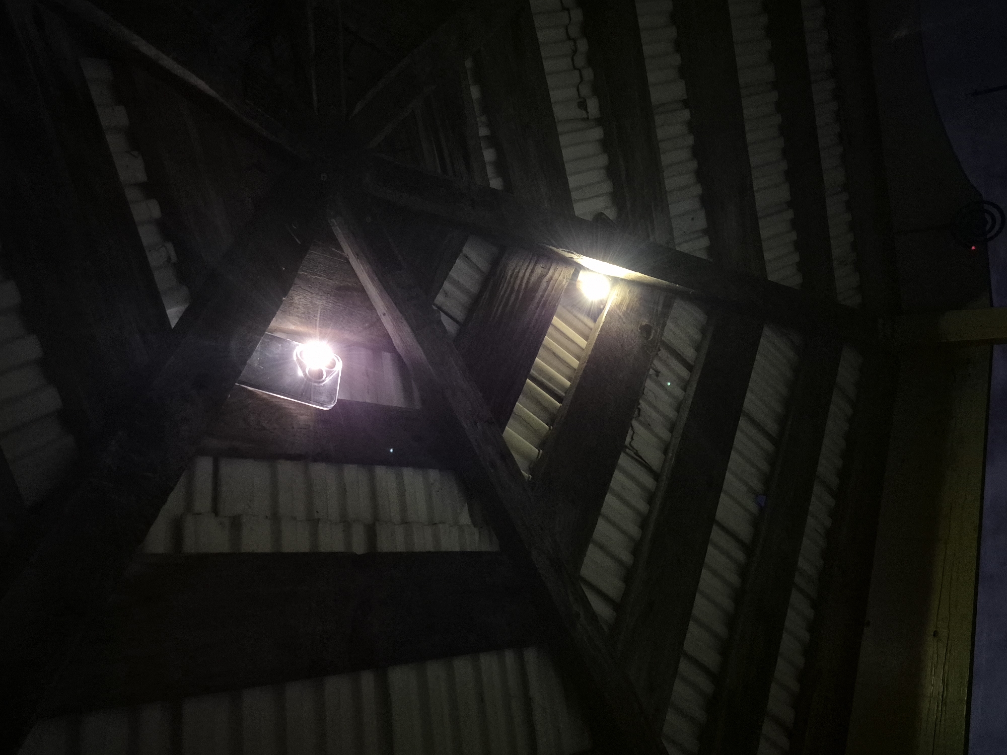 This is a low-angle, close-up, indoor shot of the roof structure of a building, likely a pavilion or gazebo, in Sars, Russia. The roof is constructed with dark wooden beams radiating from a central point and supporting corrugated metal roofing panels. Two bright light fixtures are visible, one near the top center and another lower and to the left, casting strong light and lens flare. The overall lighting is dim, with shadows obscuring much of the detail within the structure. The image does not contain any people or depict any actions or interactions.