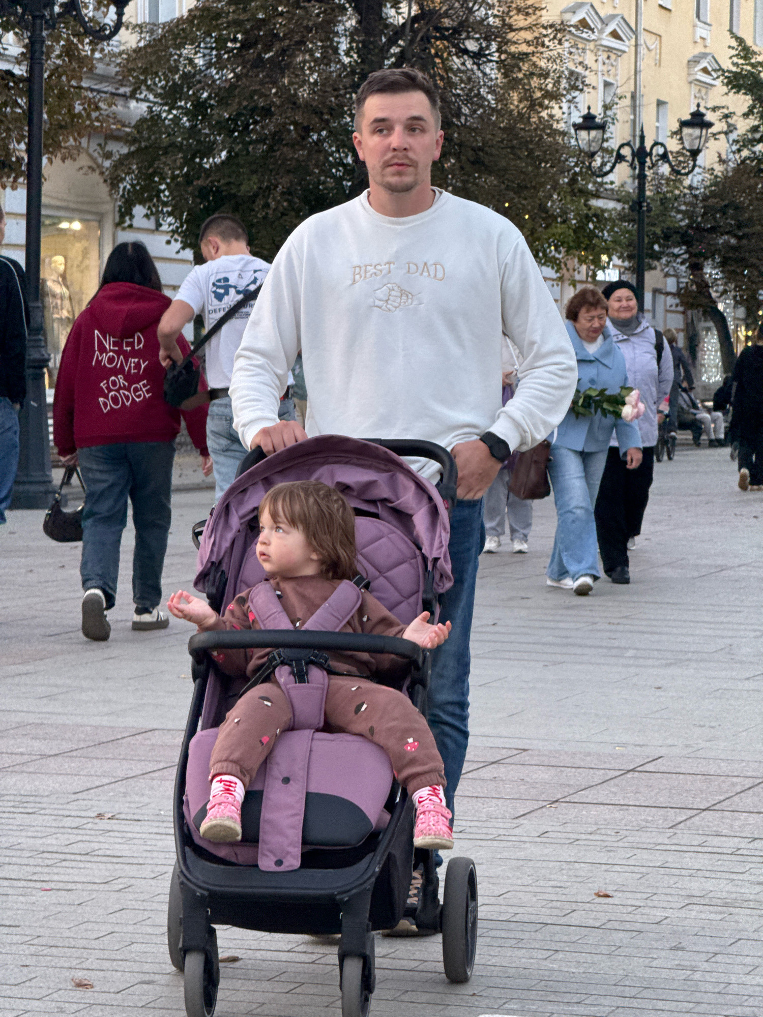 The image captures a street scene in the city of Tver, Russia, likely during autumn given the foliage and overcast sky. In the foreground, a man in a white sweatshirt with the inscription "BEST DAD" stands behind a purple stroller containing a young child. The child, dressed in a brown outfit with a pattern, is looking to the left. Behind them, other pedestrians are visible, including a person in a red hoodie with the text "NEED MONEY FOR DODGE" on the back. Further in the background, a woman holding a bouquet of pink roses walks in the same direction. The environment appears to be a public pedestrian walkway with buildings and lampposts lining the street.