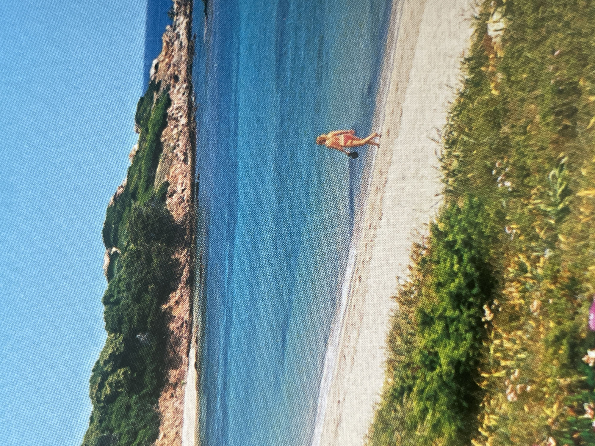 The image depicts a scenic coastal view with a person walking on a sandy beach. The sea is a vibrant blue, meeting a shoreline with a gentle wave. In the background, a lush green hillside slopes down towards the water, dotted with rocky outcrops. The sky is clear and bright blue. A solitary individual, appearing to be a woman in a reddish-orange bikini, is walking along the water's edge, carrying a dark object in her left hand. The foreground features wild grasses and some blooming flowers. The scene is an outdoor environment, likely a beach or coastal area, under daylight conditions. There is no indication of this location being in Sofia, Bulgaria; it appears to be a Mediterranean or similar coastal setting.