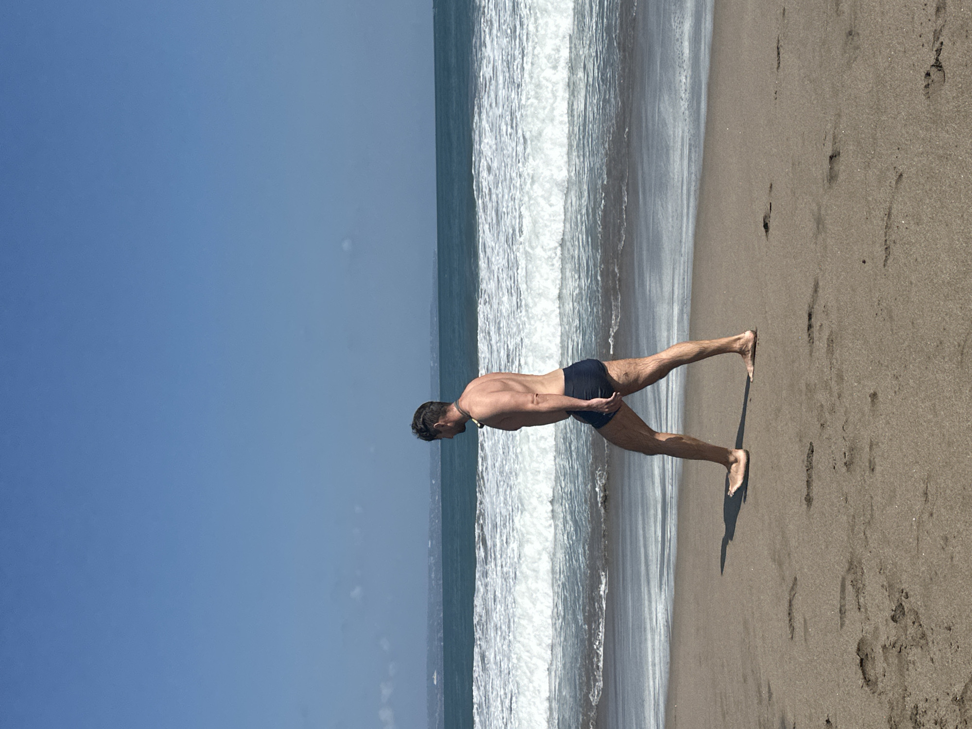 A man wearing dark blue swimming trunks walks along a sandy beach in North Kuta, Indonesia, parallel to the ocean. He is facing right in profile, with his left leg extended forward and his right leg bent. The sun casts a shadow of his legs onto the sand. The ocean is to his right, with white waves breaking and receding onto the shore. The sky above is clear and blue. The beach is brown sand with footprints visible. The man has dark hair and appears to be in his 40s or 50s. He has a light-colored object around his neck.