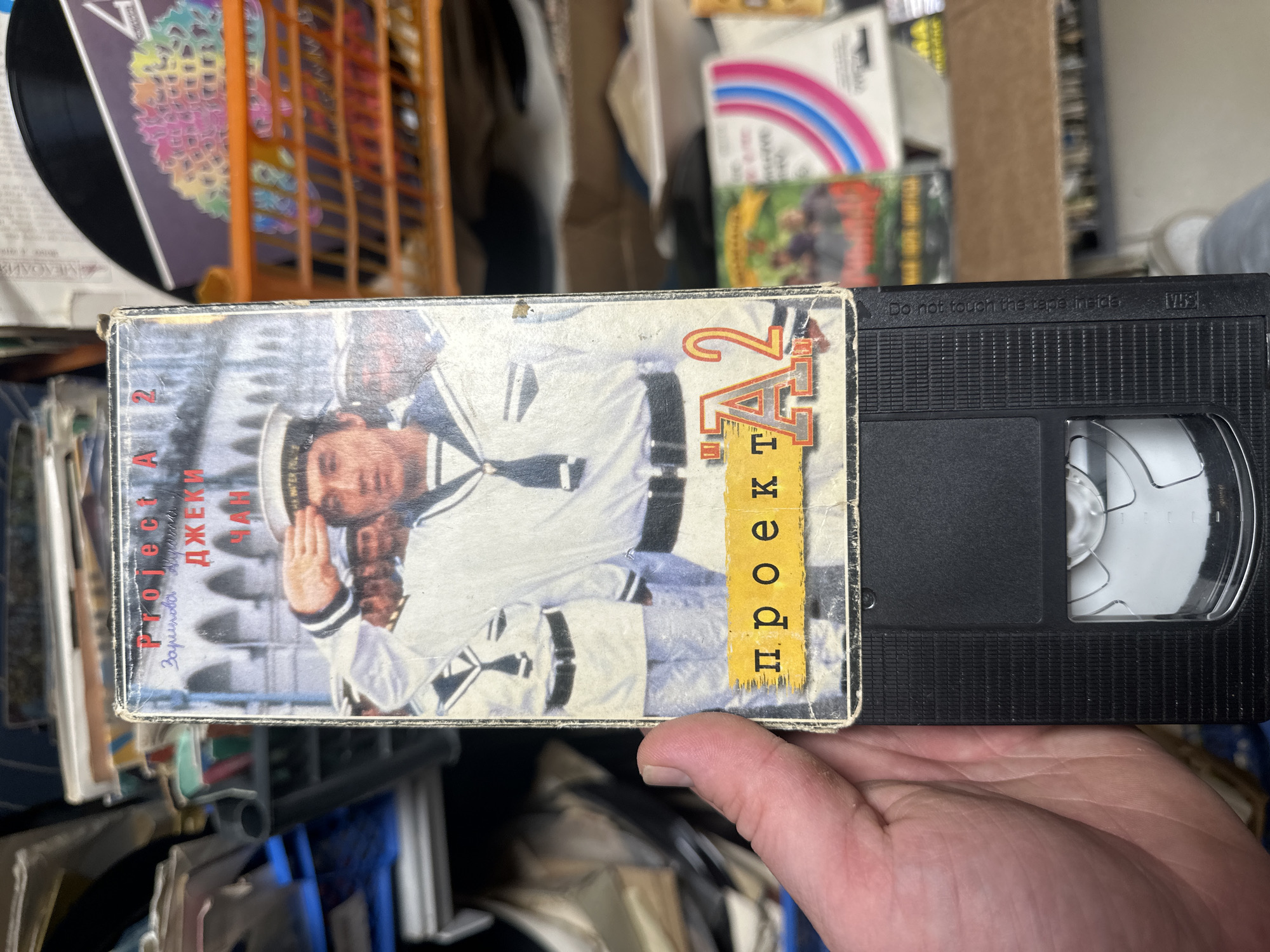 The image shows a hand holding a VHS tape of the movie "Project A 2" starring Jackie Chan. The tape is black and the cover art features Chan in a white naval uniform. The tape is surrounded by other items, including records and boxes. The location is not provided in the image.