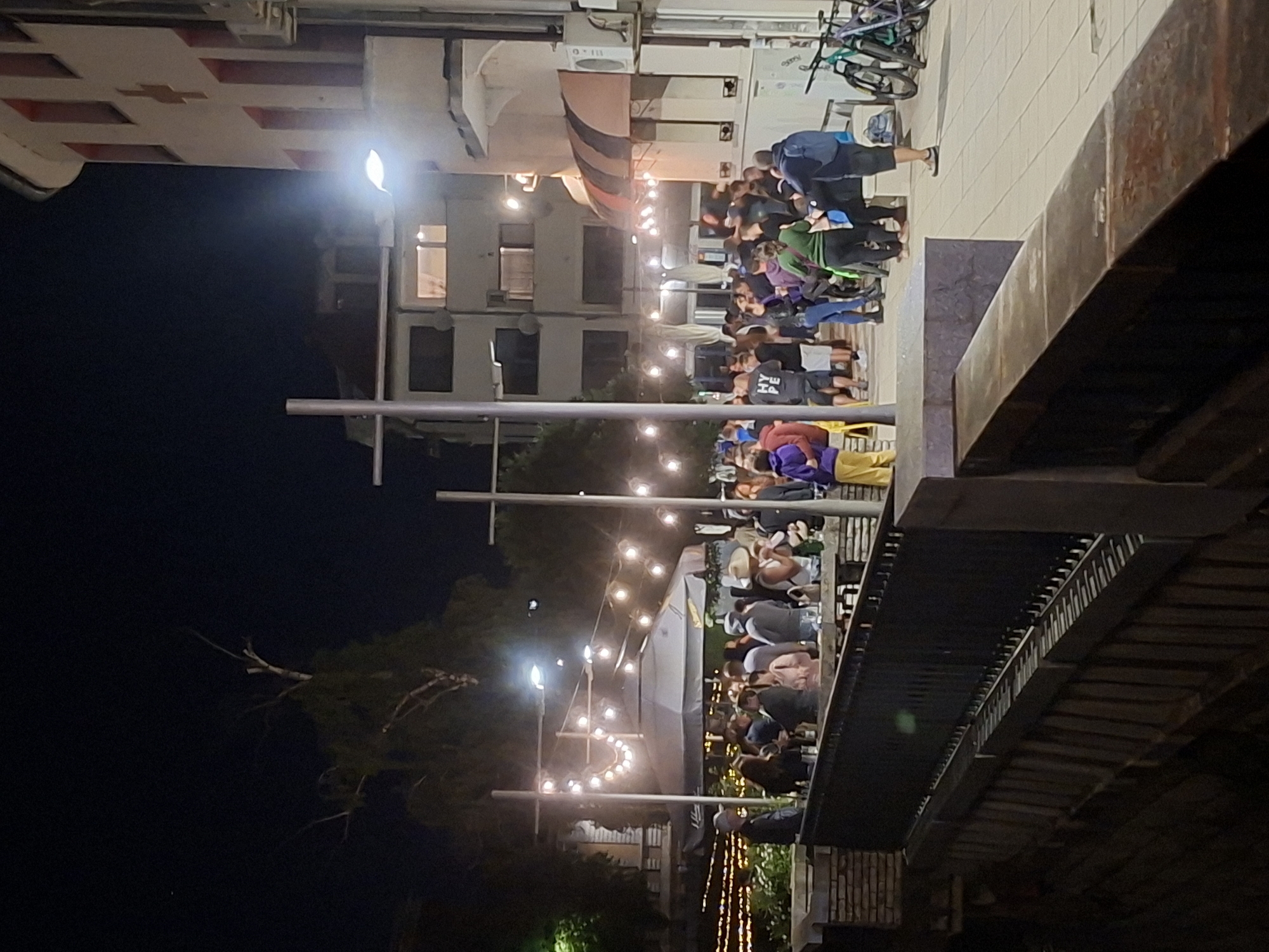 The image depicts an outdoor dining scene in Gabrovo, Bulgaria at night. Tables are filled with people, suggesting a restaurant or cafe setting. String lights are hung above, adding illumination. The setting is near a bridge and under scaffolding. Buildings are in the background.