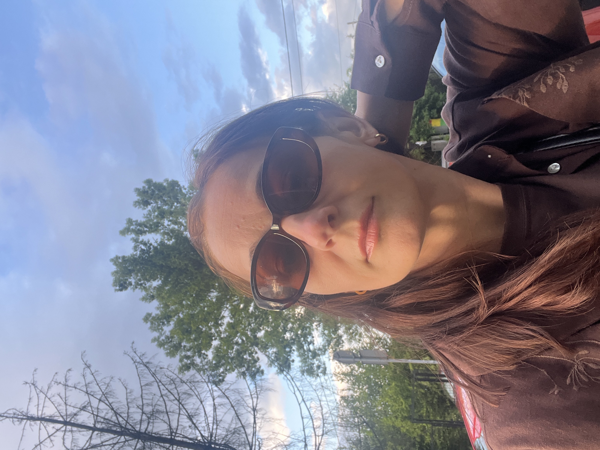 A woman is captured in a close-up outdoor selfie. She has long, reddish-brown hair parted on her left, and wears dark brown sunglasses with thick frames. Her lips are visible with a light-colored gloss. A small gold hoop earring is visible in her right ear. She wears a dark brown button-up shirt or jacket over a lighter brown undershirt. The visible left sleeve features a cuff with a silver-toned button. An embroidered floral or leaf pattern is present on the right side of her outer garment. Her left arm is raised with the hand positioned behind her head/neck area.
The background consists of a partially cloudy blue sky, with brighter blue on the right indicating the sun's direction. To the left, a tall, bare deciduous tree with dark, intricate branches stands against the sky. To the right, lush green deciduous trees with full foliage are visible. Subtle urban elements include a light-colored pole to the left, faint vehicle reflections (red) in the lower left, and overhead wires in the upper right. The scene is illuminated by natural, direct sunlight, casting highlights on the woman's face and hair, particularly on her right side. The location is an unidentified outdoor setting in the city of Unknown, Unknown.