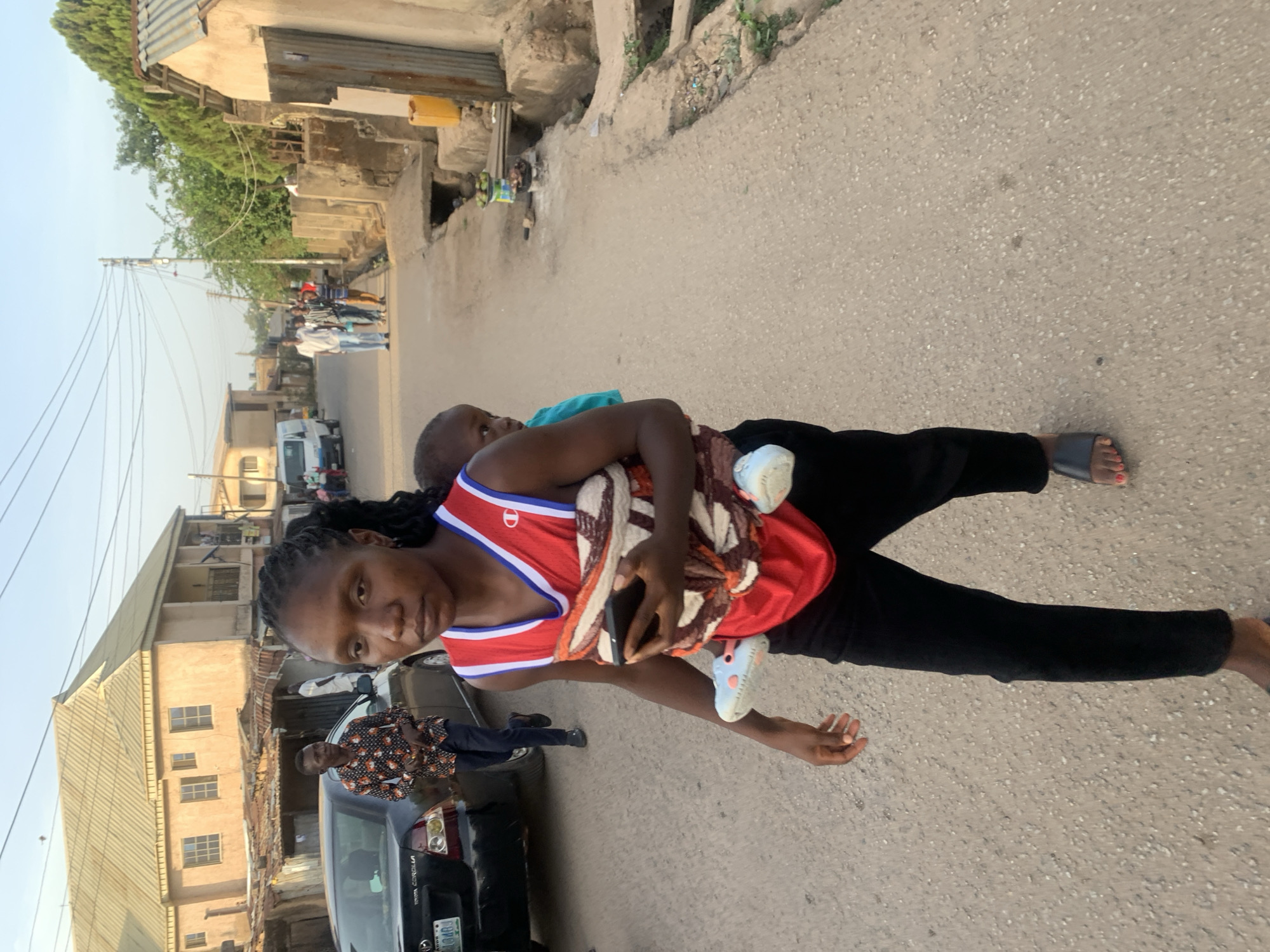 The image depicts a daytime street scene in an urban or semi-urban environment, consistent with Jos, Nigeria. In the foreground, a young woman with braided hair stands on a paved road, looking directly at the camera with a neutral expression. She is wearing a red and white athletic-style tank top with a distinctive 'D' logo and black pants, paired with dark sandals that reveal red painted toenails on her right foot. She is holding a small child, wrapped in a brown and cream patterned cloth, against her body. The child wears light blue and white infant shoes and appears calm or asleep. The woman also holds a dark mobile phone in her left hand.
Behind her, the paved road stretches into the distance, where several other individuals can be seen walking. To the left, a dark sedan, identified as a "TOYOTA COROLLA" from faint text, is parked or moving slowly. Its Nigerian license plate "LSD-104D" is partially visible. Another person in a patterned shirt stands near this vehicle. Further down the street, a white van is also discernible.
The background features various buildings, some with corrugated metal roofs and concrete structures, lining both sides of the road. Dense green foliage can be seen above some of the structures. Utility poles with numerous overhead power lines crisscross against a clear, bright sky, indicating fair weather. A yellow jerrycan is visible near a building on the right side of the road.