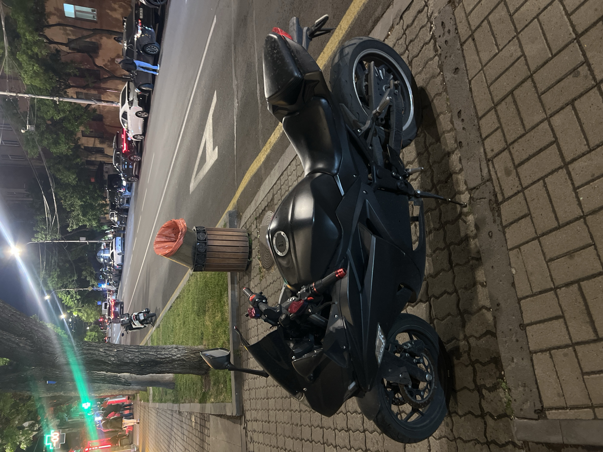 A black motorcycle is parked on a brick sidewalk in Unknown, Unknown, next to a trash can. The street adjacent to the sidewalk shows a line of parked cars. Street lights illuminate trees, buildings, and a road with vehicles. In the distance, there are more street lights and cars.