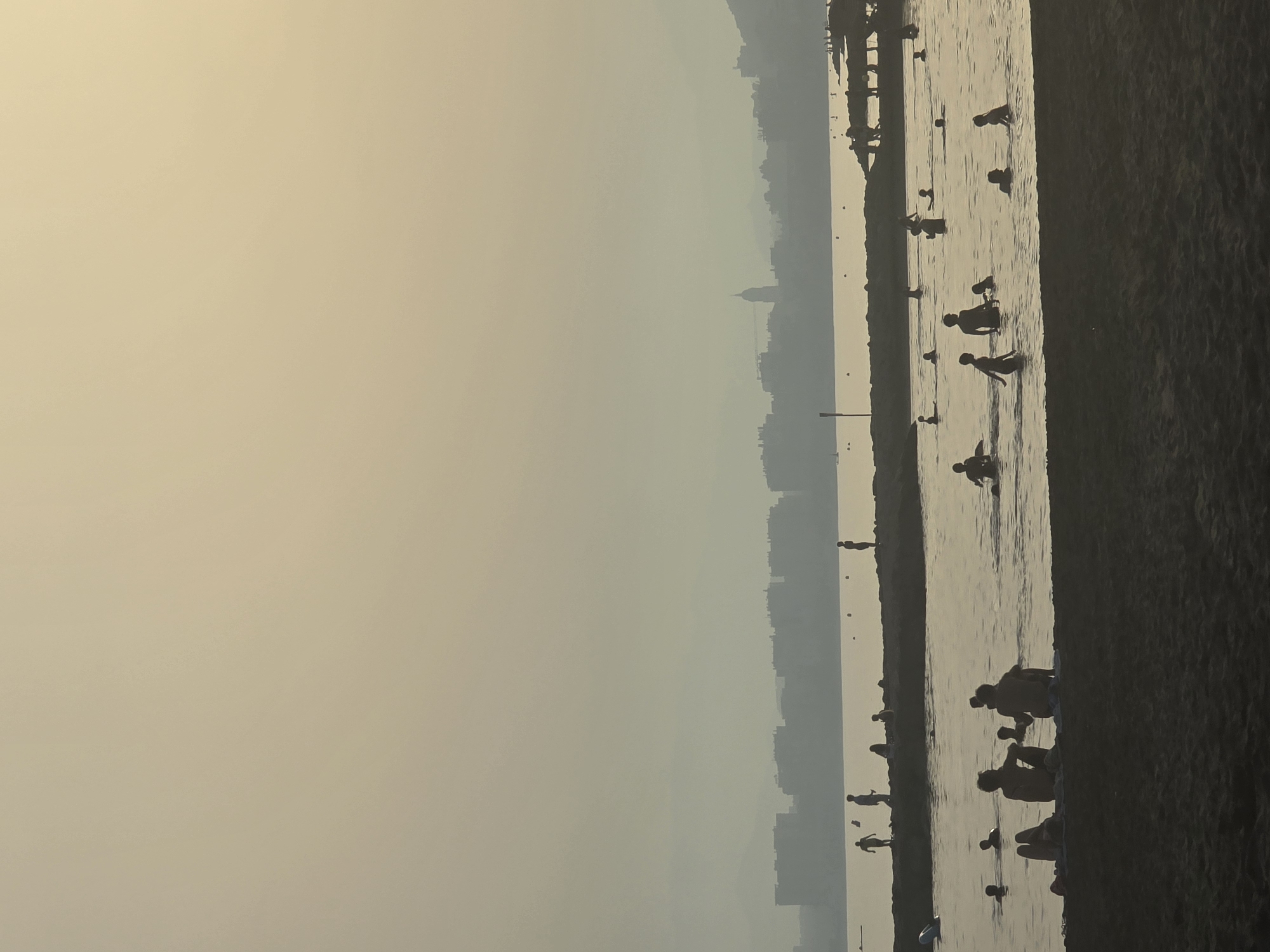 The image depicts a dusky scene at a beach in Malaga, Spain, with a hazy skyline of buildings and a church spire visible in the distance across a body of water. The foreground features a dark, silhouetted beach area with several figures sitting or lying down. Further out, a stone breakwater separates the beach from a sea populated by numerous silhouetted individuals wading, swimming, and standing in the water. Some people are interacting, while others appear to be alone. The overall atmosphere is serene and slightly muted due to the atmospheric haze and the time of day.