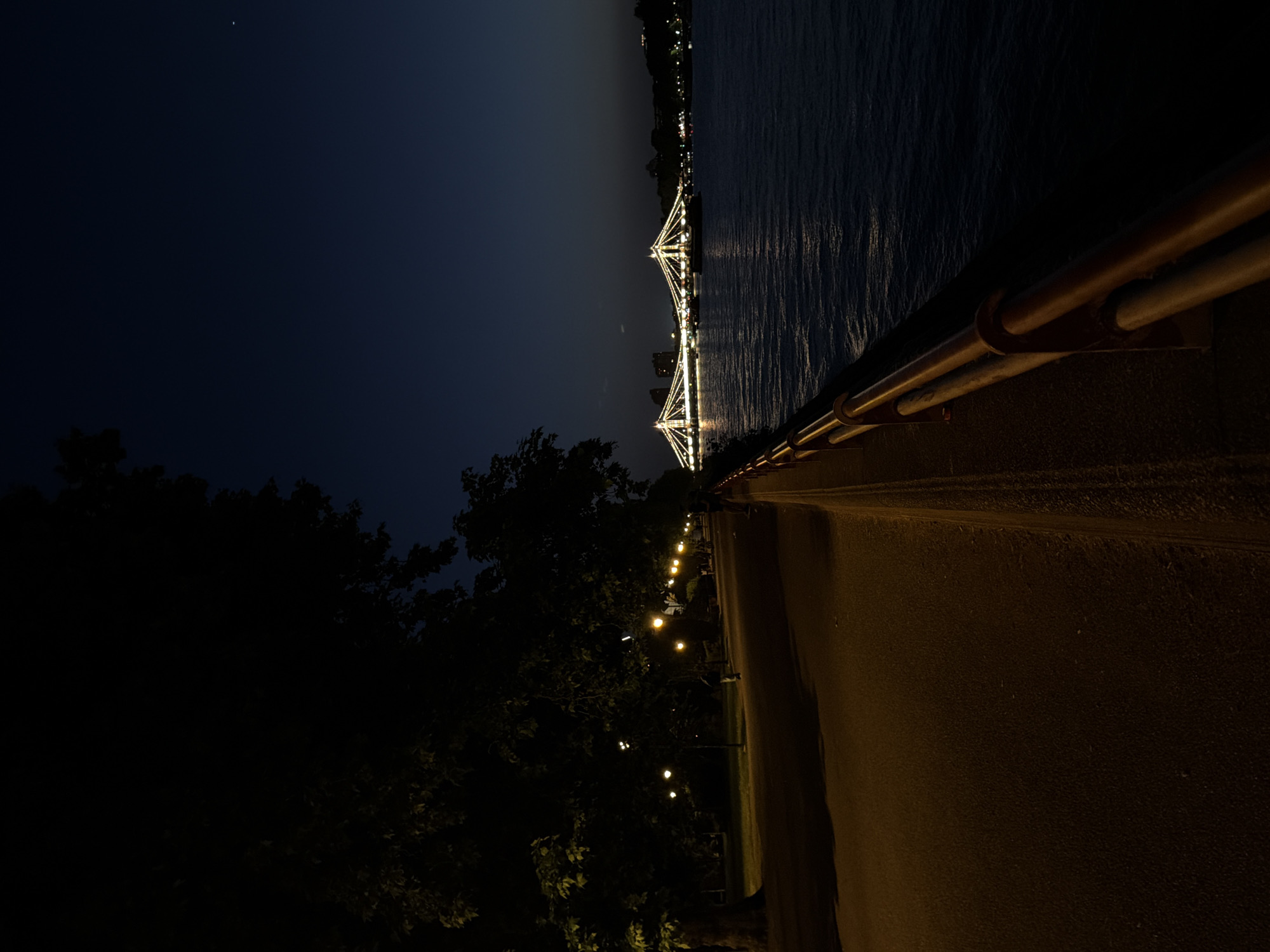 The image depicts a nighttime scene in London, United Kingdom, featuring the illuminated Hungerford Bridge spanning the River Thames. A pedestrian walkway with a railing is visible in the foreground, lined with trees and streetlights. The sky is dark, with a single visible star. The river reflects the lights of the bridge and the city.