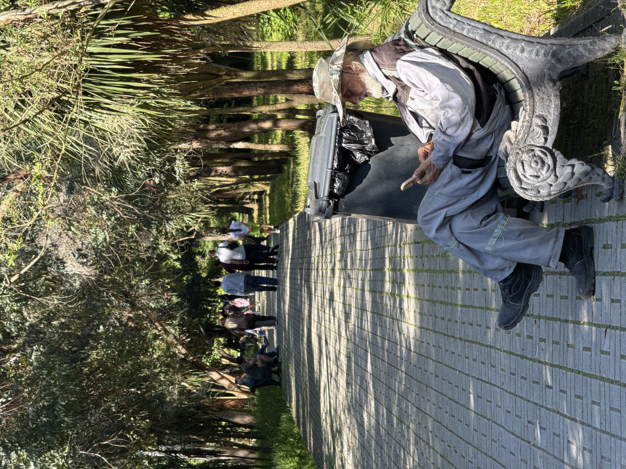 An elderly man, wearing a camouflage hat, grey overalls, black boots, and a light grey jacket, is seated on a dark grey bench. He appears to be resting his head on his hand, and his other hand holds a small, light-colored object. To his left is a dark grey trash receptacle with a black plastic bag inside. The bench is situated on a paved walkway composed of grey rectangular pavers with green grass growing between them. A group of people can be seen walking in the distance along the walkway. The location is surrounded by greenery, including trees and shrubs. The city, Sirius, Russia, is not discernable from the image alone.