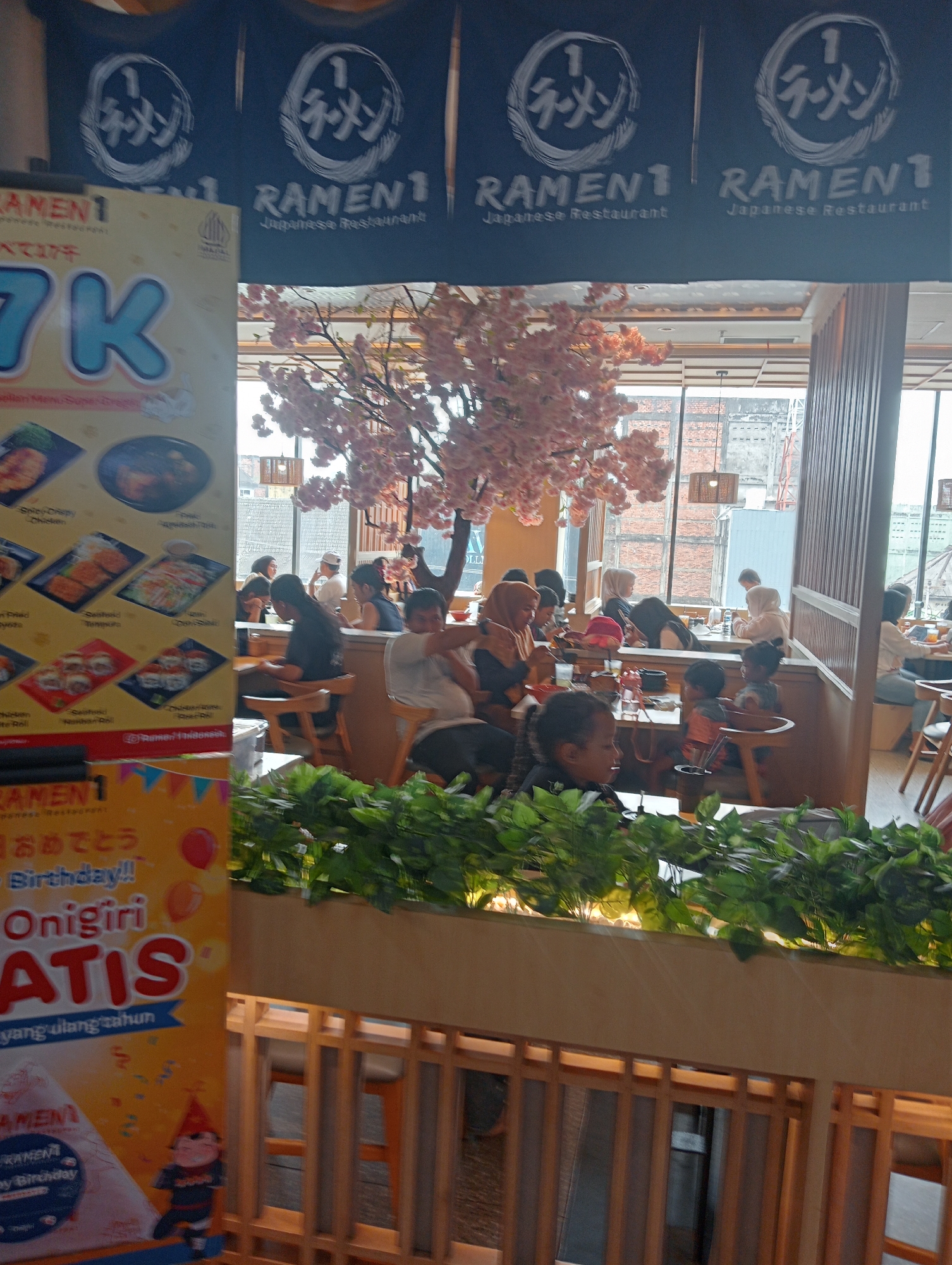 The image depicts the interior of a Ramen 1 restaurant, likely located in Jambi City, Indonesia, as indicated by promotional materials in Indonesian language. The restaurant has a modern and inviting ambiance, with large windows offering a view of the city outside. Inside, patrons are seen enjoying their meals at tables, with some engaged in conversation while others focus on their food. The decor features elements of Japanese culture, such as a decorative cherry blossom tree and banners with Japanese characters and the Ramen 1 logo. The lighting appears to be a mix of natural light from the windows and artificial lighting, creating a warm and cozy atmosphere. The scene is lively with a moderate number of people present, suggesting a busy lunch or dinner service.