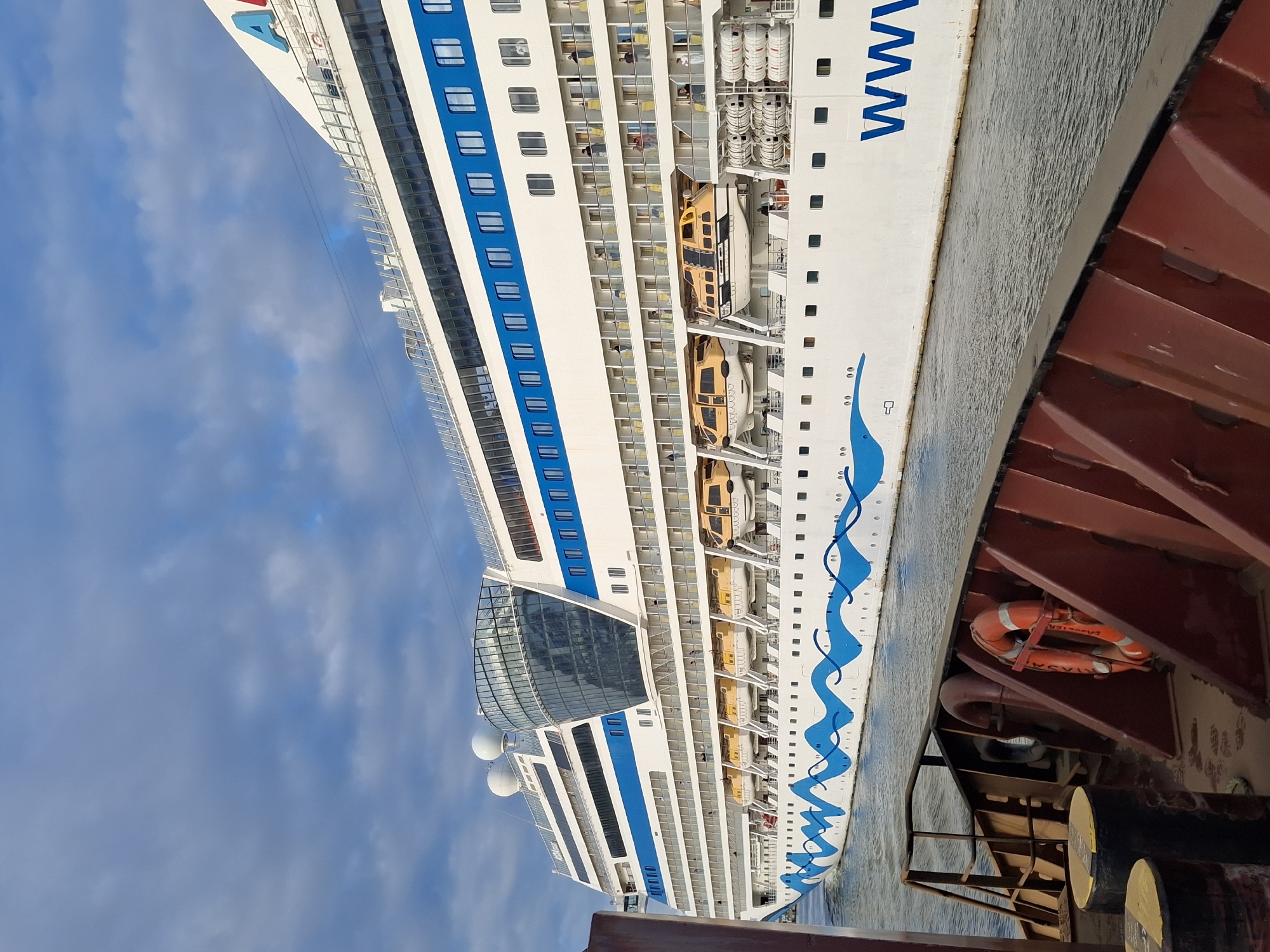 The image captures a partial view of the cruise ship AIDAmar, docked in Rostock, Germany. The ship's hull is predominantly white, adorned with horizontal blue stripes and a distinctive blue wave pattern near the waterline. Several rows of windows are visible, suggesting multiple decks with passenger cabins. Lifeboats are visible along the side. The ship is docked alongside a red structure, with a life preserver attached to its interior. The sky is visible above the ship, featuring a mix of blue and white clouds. Several large, cylindrical items are seen at the bottom of the frame.