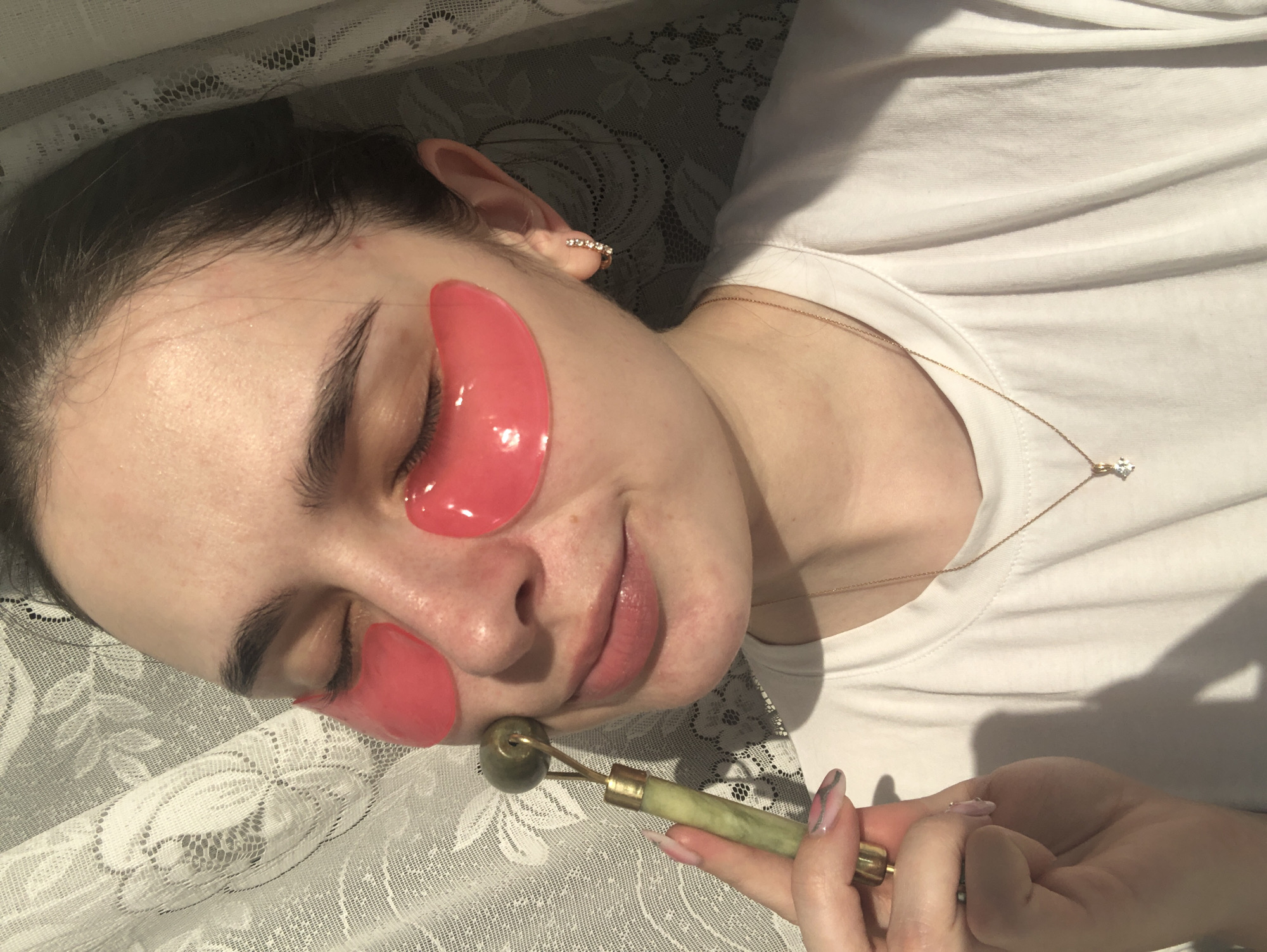 The image captures a young woman from Noviy, Russia, engaged in a self-care beauty routine indoors. She is seen lying down, with her eyes closed and a subtle, relaxed smile on her face. Her dark hair is pulled back, revealing her forehead, which has a small blemish.
The primary subjects are the woman and the items she is using for her skincare. She has vibrant pink, translucent gel eye patches applied under both eyes. In her right hand, she holds a jade roller, characterized by its green stone and gold-colored metal handle, which she is gently applying to her jawline or chin area. Her fingernails are manicured with a pink and clear gradient design.
She is wearing a simple white t-shirt and accessorized with a delicate gold chain necklace featuring a single, clear, square-cut stone pendant. A small, sparkling ear cuff or earring is visible on her right ear.
The setting appears to be a comfortable indoor space, possibly a bedroom, indicated by the white lace-patterned fabric visible in the foreground and background, which could be a bedspread, sheet, or curtain. Bright, natural light illuminates her face and upper body, suggesting the time of day is daytime. The overall mood is one of relaxation and personal care. There is no visible text in the image.
