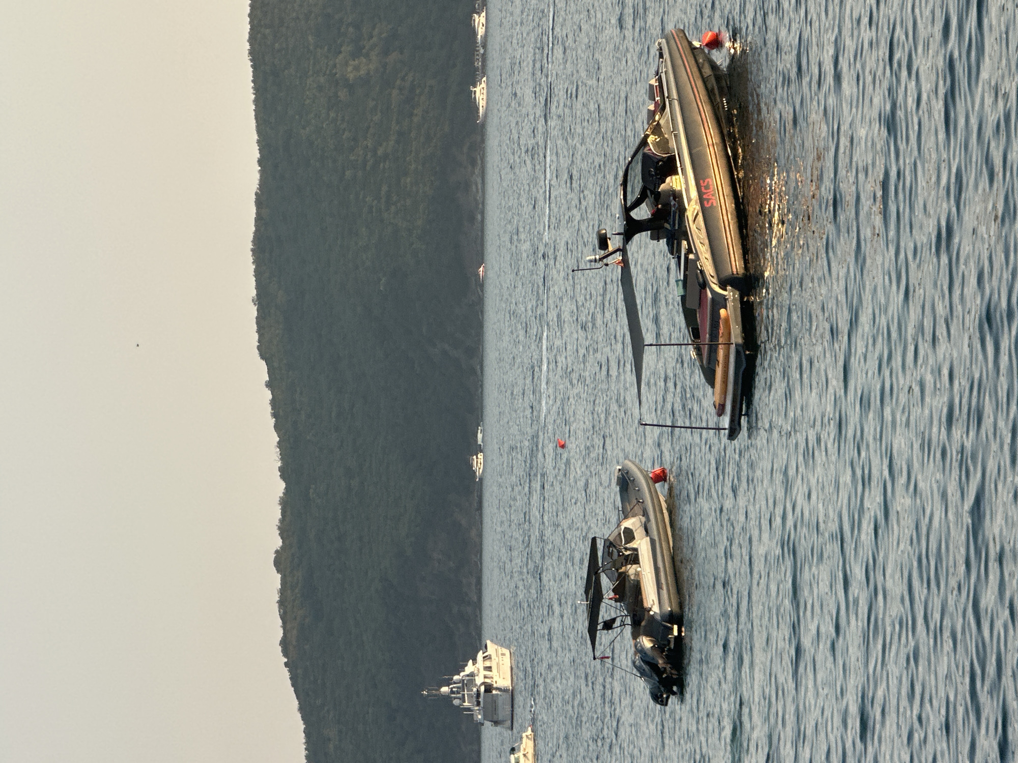 The image depicts a calm sea with several boats anchored or cruising. In the foreground, two inflatable speedboats are visible. The boat on the right is larger, dark gray with orange trim, and has "SACS" written in red on its side. It is equipped with a canopy, a steering console, and seating. A person is seated at the console. A small orange buoy is attached to the side of this boat. The boat on the left is also an inflatable speedboat, lighter gray, with twin outboard motors at the stern. It has a canopy and a person is visible in the boat. Another orange buoy is attached to this boat.
In the mid-ground, a large white motor yacht with multiple decks and a helipad is visible. Further in the distance, other smaller boats and yachts are scattered across the water. The background features a densely vegetated, dark green hill or mountain range, characteristic of the Budva Riviera in Montenegro. The sky is clear and a pale yellow, suggesting either early morning or late afternoon light. The water is calm with gentle ripples. The overall scene conveys a sense of leisure and maritime activity in a coastal environment.