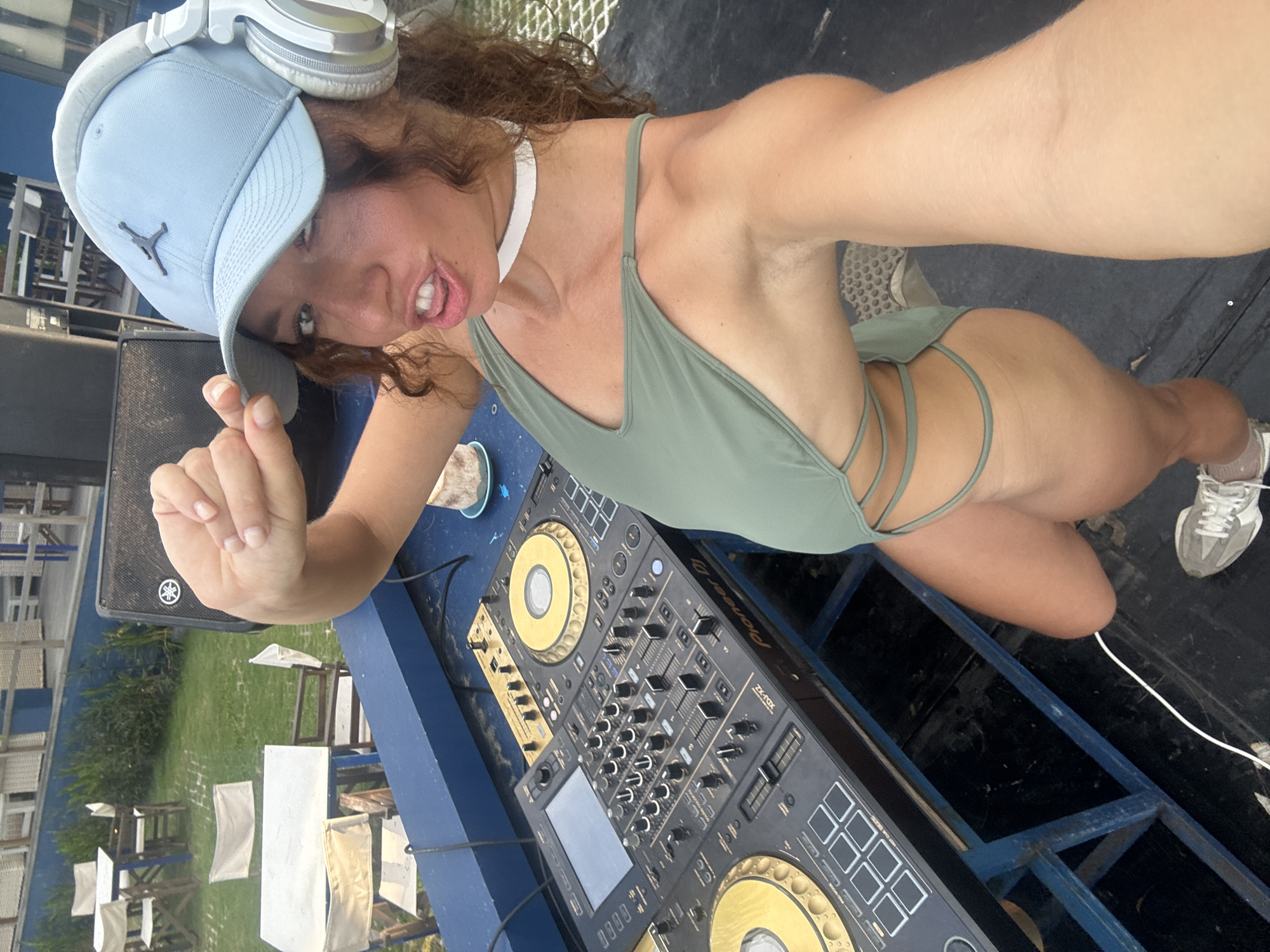 A tanned woman in a green swimsuit and white sneakers is taking a selfie. She is kneeling next to a DJ mixing console, wearing headphones and a light blue Jordan baseball cap. The console is black and gold. A Yamaha speaker is to her left. White chairs and tables are visible in the background, suggesting an outdoor venue. The location appears to be Đà Nẵng, Vietnam.
