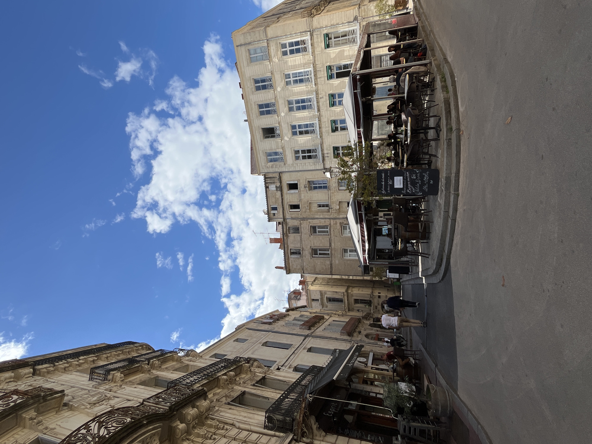 The image depicts a street scene in Montpellier, France, characterized by tall, weathered buildings with multiple windows and ornate wrought-iron balconies. The sky is bright blue with scattered white clouds. Along the street, outdoor seating areas of cafes or restaurants are visible, furnished with tables and chairs. Some people are walking on the pavement. The street is paved and shows signs of use. Chalkboard menus or signs are also present.