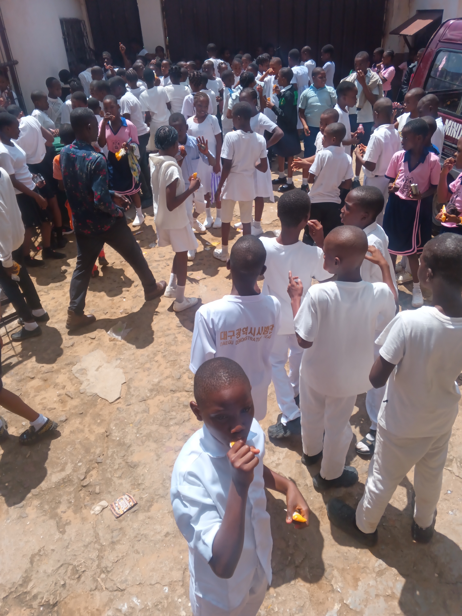 In Onitsha, Nigeria, a group of young students in white school uniforms gather outdoors in a dusty, uneven courtyard. The scene appears to be in front of a building with a dark, large doorway, possibly a school or community center. Several children are holding and eating snacks, suggesting a break or a special event. A portion of a maroon van with "MIND..." visible on its side is present on the right side of the frame, indicating a potential mode of transport or service. The ground is a mix of dry soil and patches of concrete or stone. The overall atmosphere seems energetic and somewhat chaotic, with children engaged in various activities.