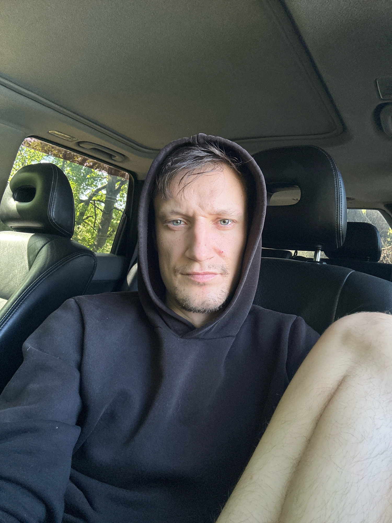 A young adult male with fair skin, light brown hair, and green eyes is captured in a selfie from the chest up, seated inside a vehicle. He is wearing a black hooded sweatshirt with the hood pulled up. The interior of the vehicle is visible, including black leather seats and a sunroof. Sunlight streams through the windows, illuminating the scene and casting shadows. Outside the vehicle, green foliage from trees is visible. The man's expression is neutral, and his gaze is directed at the camera. His right leg is visible in the foreground, with visible hair on the skin. The overall impression is that of a casual, personal snapshot taken outdoors. The image is not geo-tagged, therefore, its location in Makiv, Ukraine cannot be confirmed.