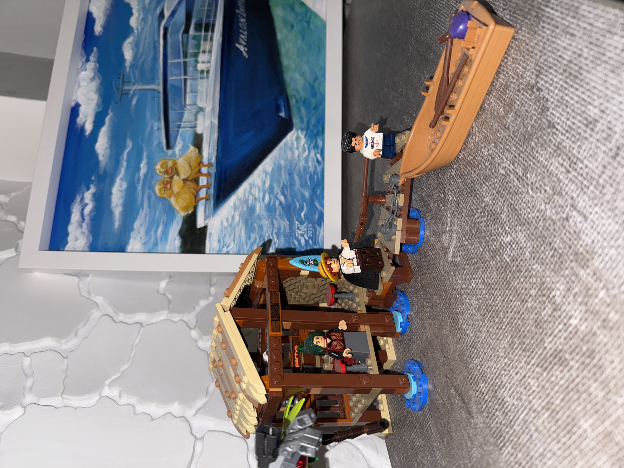 The image displays a playful arrangement of Lego sets against a domestic background, with a framed painting on the wall. The primary subjects are several Lego minifigures and two Lego structures.
On the right, a Lego minifigure with dark hair and a white shirt featuring the word "ANCHOR" stands beside a small, light brown Lego dinghy containing two brown oars and a purple rounded piece. This dinghy is connected to a larger, brown, multi-level Lego structure, possibly a raft or a section of a ship, which rests on a textured gray surface. This main structure has a roof, intricate brown detailing, and blue circular pieces at its base. Two other Lego minifigures are positioned on this structure: one with a straw hat, white shirt, and dark jacket, holding a blue surfboard, and another with green hair and a red and black outfit, standing inside the structure. A small, dark gray cannon-like piece is attached to the front of this structure.
In the background, a framed painting depicts a large blue ship with the name "Avalon Div..." partially visible on its side, sailing on blue water under a sky with white clouds. Two yellow ducklings are depicted in the foreground of the water. The painting also shows what appears to be a signature with "SK" and the year "2015". The wall behind the Lego setup features a white, raised, irregular pattern, while the Lego sets are placed on a gray, coarse-textured surface.
The scene appears to be an indoor setup of Lego toys, creating a fantasy or adventure narrative with the figures and structures. No specific event is actively taking place, but the arrangement suggests preparation for or an aftermath of an adventure. The time of day in the painting is bright and clear. Visible text includes "ANCHOR" on a minifigure's shirt, "Avalon Div..." and "2015" on the painting, and a partial word "partyv" on the main Lego structure.