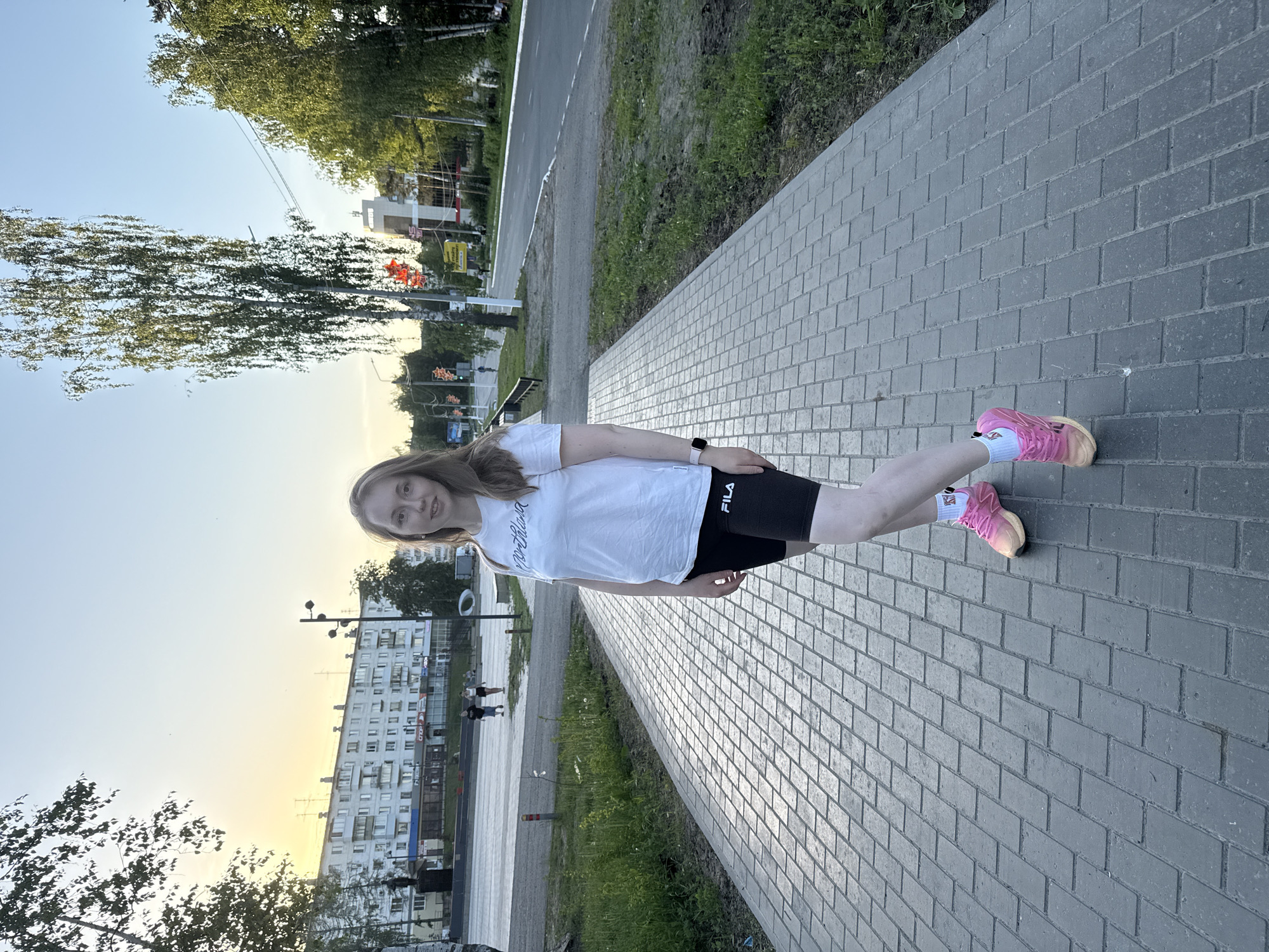 A young woman stands on a gray brick sidewalk in an urban area of Unknown, Unknown. She wears a white t-shirt, black athletic shorts, pink sneakers, and white socks. A smartwatch is visible on her left wrist. Behind her is a green grassy area separating the sidewalk from a street and buildings. Overhead power lines are visible. The buildings appear to be multi-story apartment complexes. The sky is light blue with a yellow-orange glow near the horizon, suggesting sunrise or sunset.