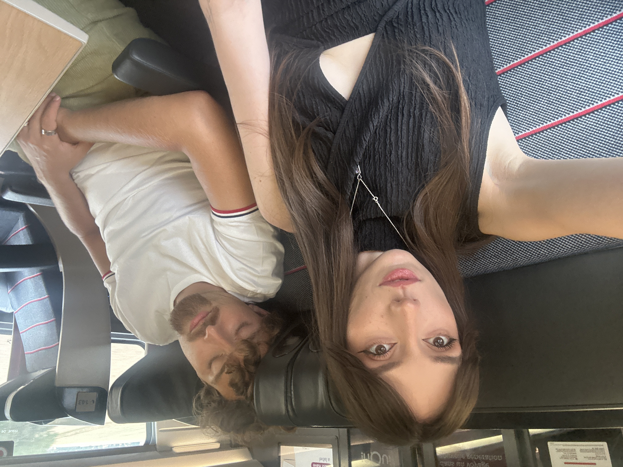 The image shows a close-up selfie of a young woman and a young man on a train. The woman is in the foreground, looking at the camera with her mouth slightly open. She has long, brown hair and is wearing a black dress with a cutout. The man is sleeping in the background, his head resting on the seat. He has brown hair and a beard and is wearing a white t-shirt. The train seats are visible, with a table to the right. The image appears to be taken during the day, with light coming from the window. The train is likely traveling through the countryside, as indicated by the blurred greenery visible outside the window. The location is specified as being in Villognon, France.