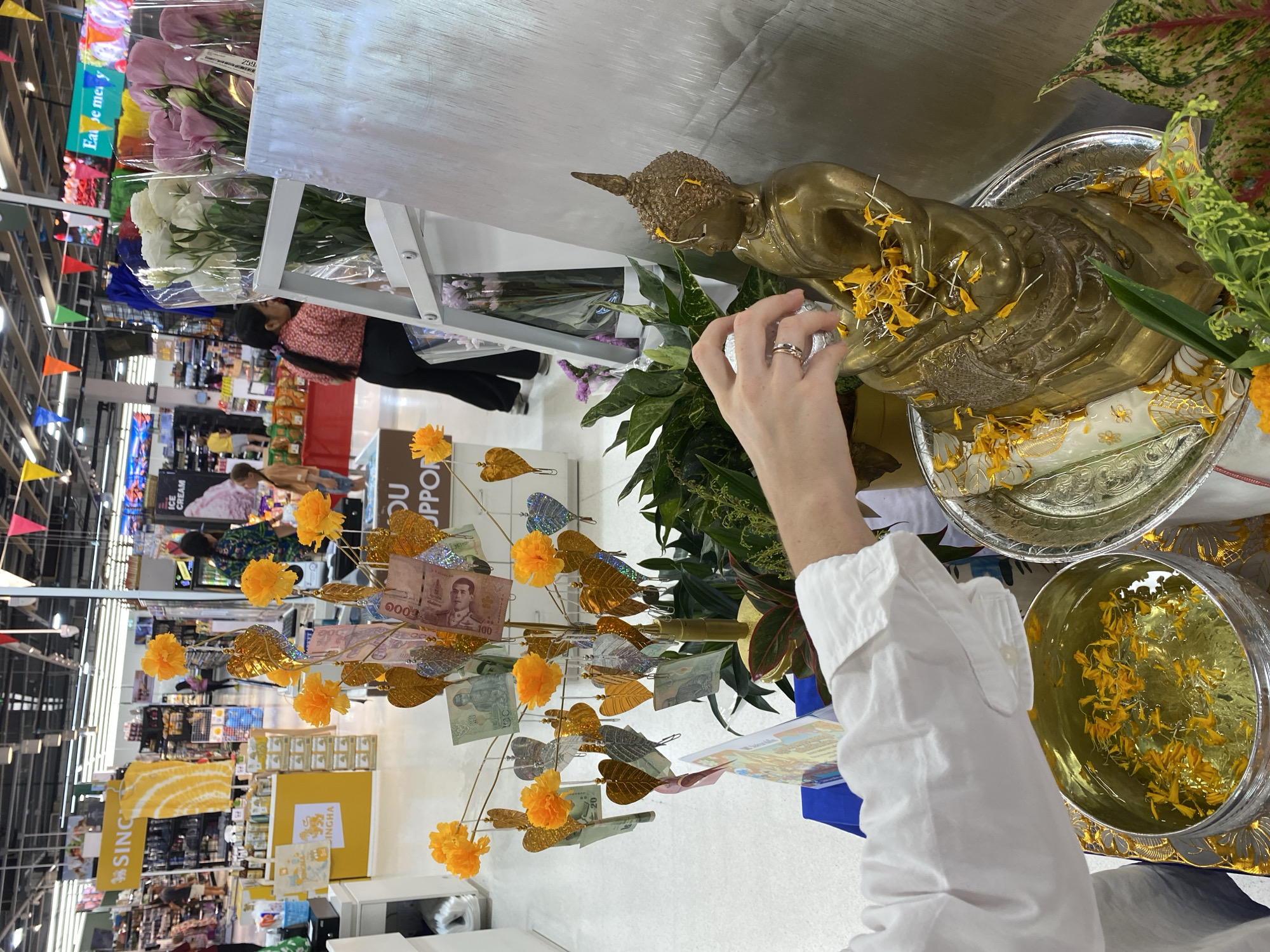 A vertical, medium shot shows a person pouring water over a gold-colored Buddha statue in a store. The person is wearing a long-sleeved white shirt. They are seen from the shoulders down and are partially in the middle right foreground. The Buddha statue is in the upper middle right. To the left of the Buddha is a tree adorned with Thai bank notes and gold colored leaves. The background of the shot is a blurred view of the interior of a store. Banners and other displays are seen in the shot. The location is Choeng Thale, Thailand.