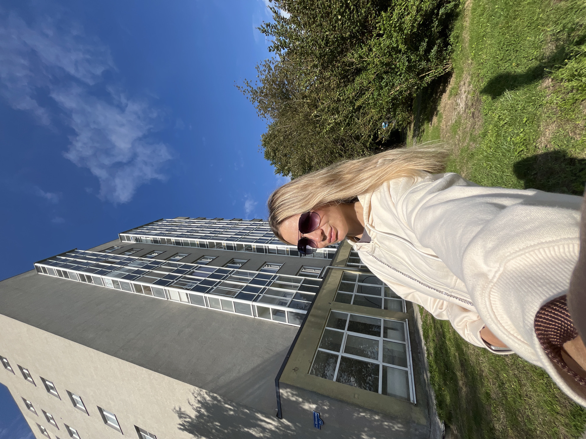 This media file is a selfie taken by a young woman with blonde hair, wearing sunglasses and a cream-colored hoodie. She is outdoors, with a tall, gray building with many windows to her left and a grassy area with trees to her right. The sky is blue with some scattered white clouds. The sun appears to be shining, casting shadows on the ground, suggesting it is daytime. The building's architecture suggests an urban or possibly institutional setting, typical of a residential area or a university campus. The overall mood is casual and relaxed, as indicated by the selfie format and the woman's attire. There is a street sign visible on the building, but the text is too small to be read clearly. The orientation of the image is rotated, making the horizon appear vertical. The location is contextually specified as Pionersky, Russia.