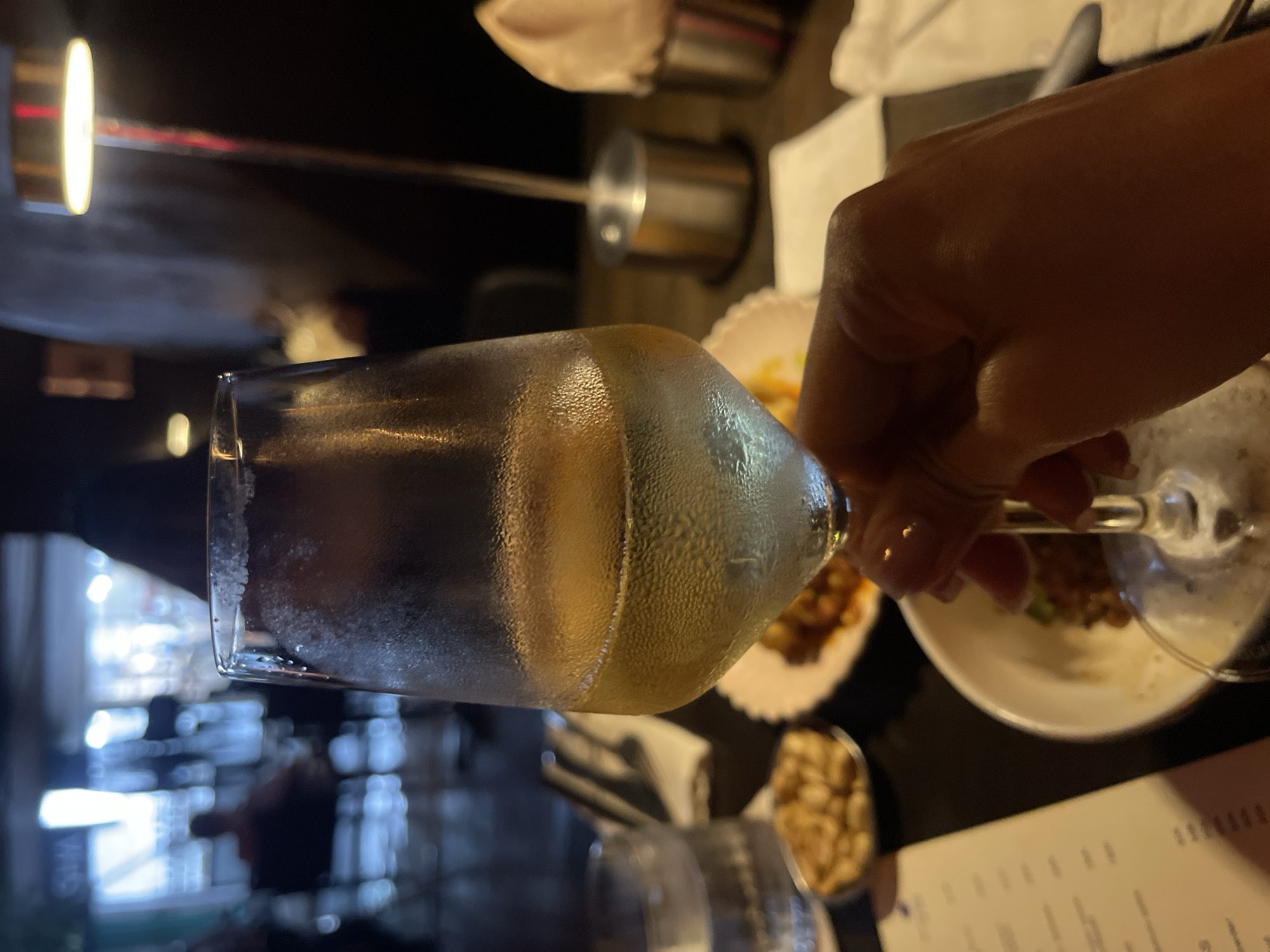 In this image, a person is holding a clear glass filled with a pale yellow liquid, possibly wine or a cocktail. The glass is condensation-covered, suggesting it contains a cold beverage. The hand holding the glass is light-skinned with manicured nails. The background is dimly lit and appears to be a restaurant or bar setting. There are out-of-focus elements in the background, including what look like plates of food, a metal container, and possibly other patrons. The overall atmosphere seems to be casual and social. The lighting suggests it is evening or indoors in a dimly lit establishment. There is no visible text in this image.