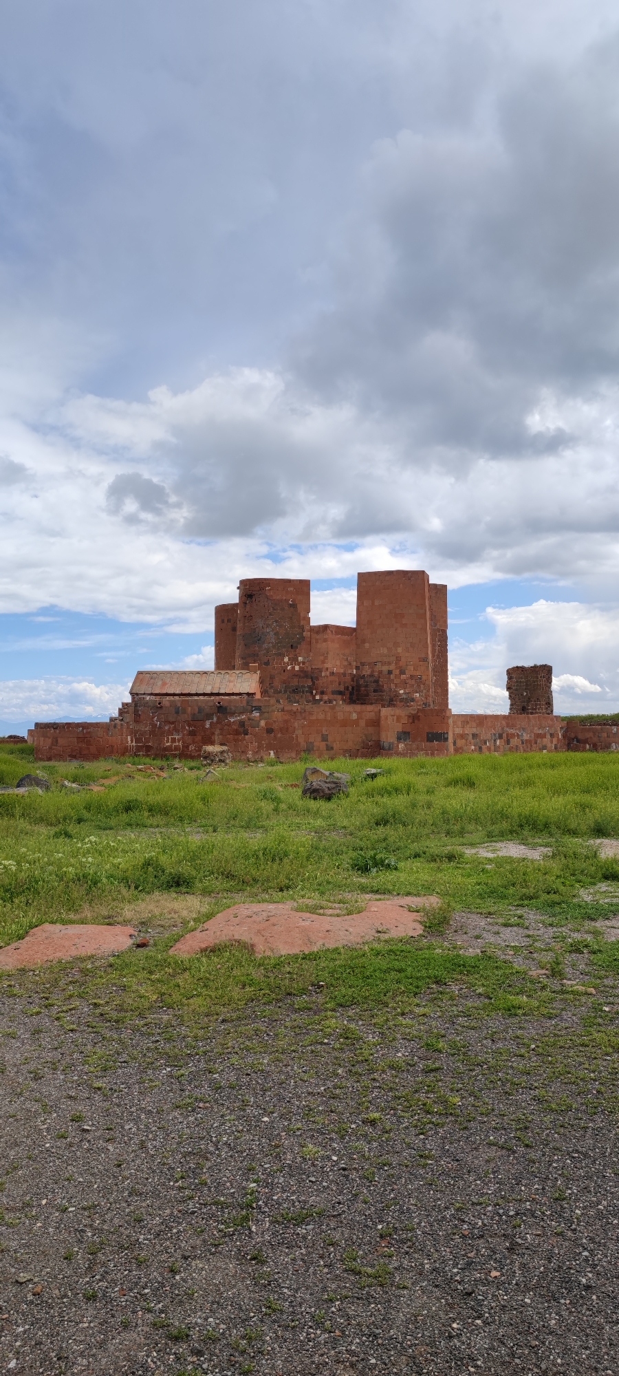 The image shows the Dashtadem Fortress in Armenia, situated against a backdrop of a partly cloudy sky. The fortress, constructed of reddish-brown stone, comprises multiple connected structures, some taller and more prominent than others. One section features a sloping roof. The structure is partially dilapidated.
The foreground is dominated by gravel and patches of green grass intermingled with red, rocky soil. The vegetation appears sparse and uneven.
