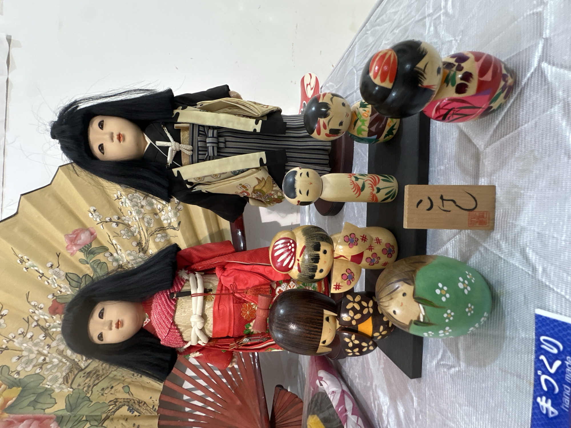 The image depicts two Japanese dolls in traditional attire and several Kokeshi dolls arranged on a surface covered with a light-colored fabric, possibly in a domestic setting, perhaps a retail store or display. The foreground features a red fan and another floral fan, which seems to be a prop. The dolls and fans suggest a Japanese cultural theme. The scene includes a small wooden plaque with Japanese characters on it. Based on the presence of Japanese dolls and related cultural artifacts, it does not appear to be in Quetta City Tehsil, Pakistan, as Japanese dolls are not commonly associated with that area.