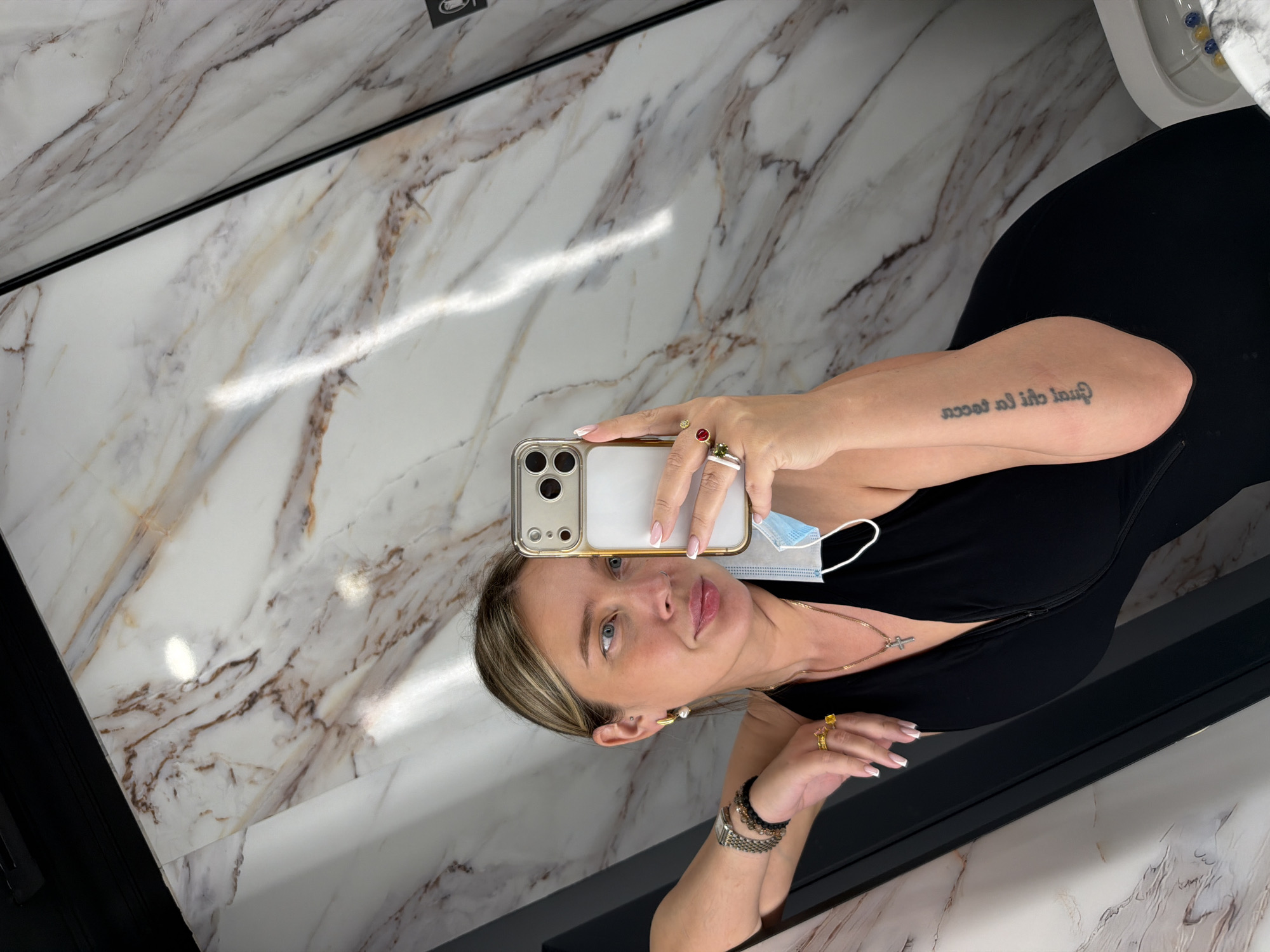A woman is taking a selfie in a bathroom with marble walls. She is wearing a black dress, jewelry, and a face mask is hanging from her ear. The location is unknown and is not necessarily in Podolsk, Russia.