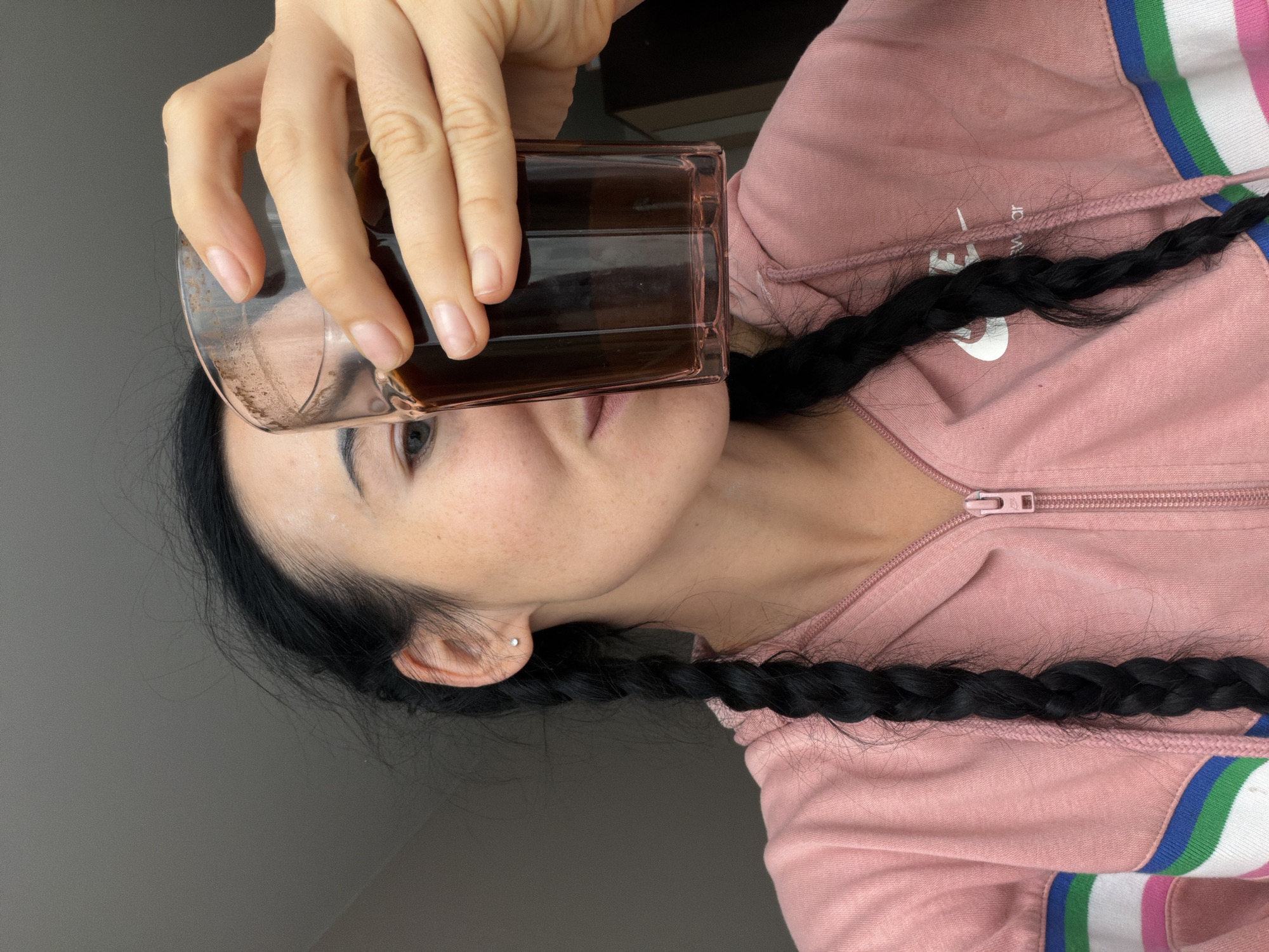 A woman with dark hair styled in two braids is holding a rectangular glass filled with a dark liquid, possibly coffee, up to her mouth. She is wearing a light pink hoodie with blue, green, and white stripes along the shoulders and sleeves. Her face is visible behind the glass, and a small earring is noticeable in her ear. The background is a plain gray wall. There is no indication of the city of Saint Petersburg, Russia, or its location.