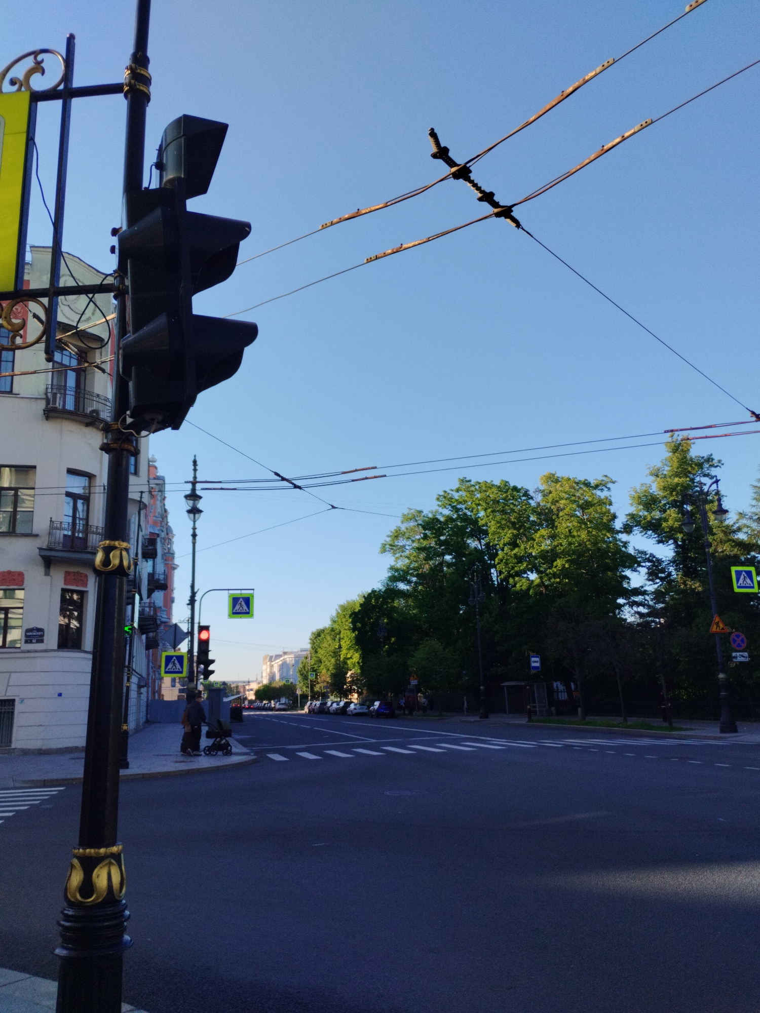 This image captures a street scene in Saint Petersburg, Russia, under a clear blue sky, suggesting it is daytime, likely morning or late afternoon given the low angle of the sun casting long shadows. The foreground is dominated by a tall, black lamppost with ornate gold detailing. Attached to it is a traffic light, currently showing red.
To the left of the lamppost, a building with classical architecture, featuring balconies and many windows, is visible. Further down the street, more buildings with similar architectural styles line the road, some adorned with red and cream facades.
Across the street, a pedestrian crossing is marked by white stripes. A pedestrian signal displaying a white walking figure on a blue background is present, along with a traffic signal showing red. A person is standing near a stroller at the corner, waiting to cross.
The street itself is dark asphalt, with cars parked along the right side of the road in the distance. Large, mature trees with lush green foliage line the opposite side of the street, creating a leafy canopy. Overhead, electrical wires, likely for trams or trolleybuses, crisscross the sky, supported by poles.
A yellow sign with some text is partially visible on the lamppost in the foreground, but the text is not legible. Other street signs, including pedestrian crossing signs, are visible. The overall atmosphere appears calm and orderly, with a hint of the urban charm characteristic of Saint Petersburg.