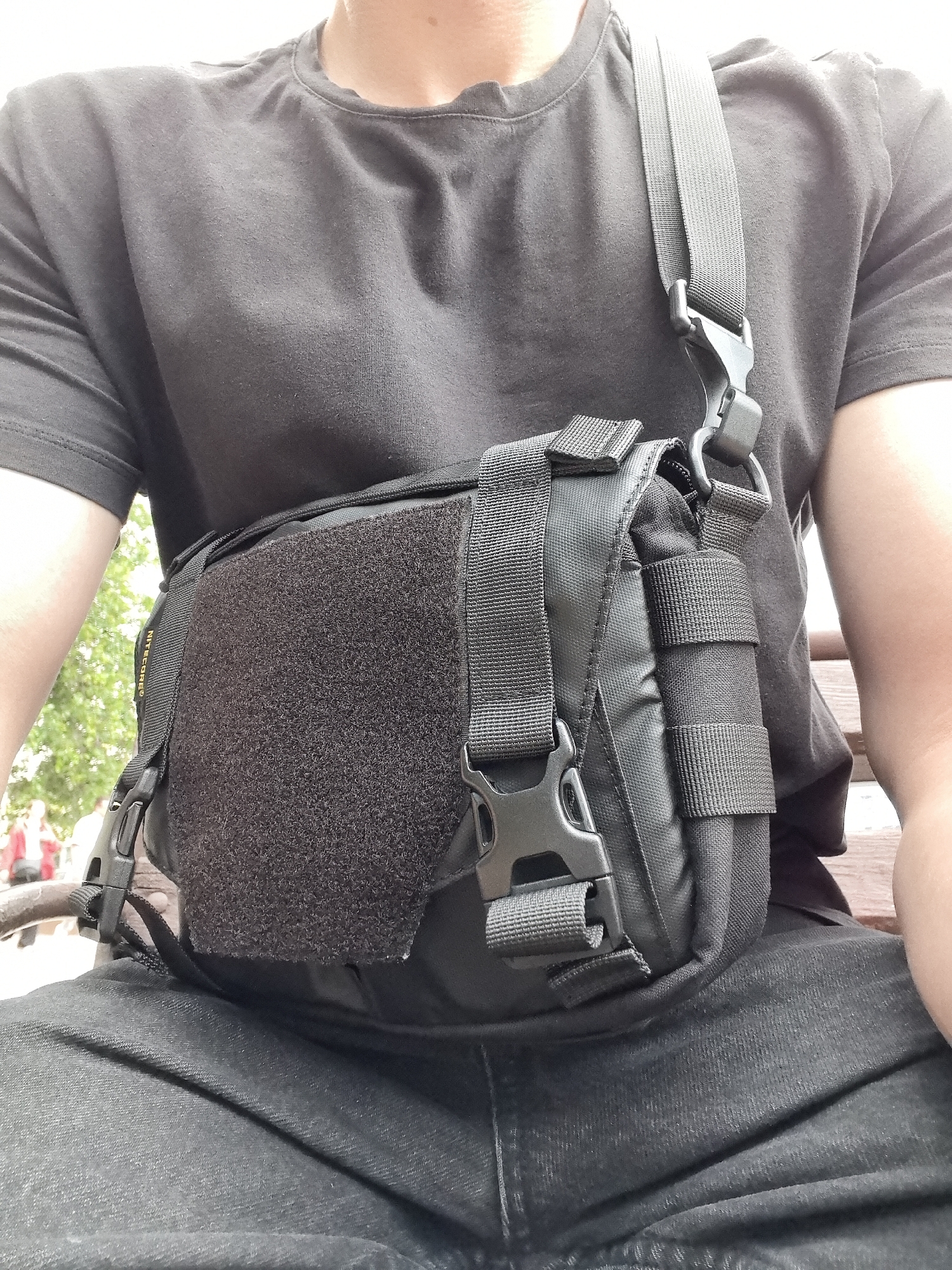 A person wearing a black t-shirt and black pants is sitting on a wooden bench outdoors. A black tactical sling bag is worn across their chest, with a strap going over their right shoulder. The bag has multiple compartments and straps, including a large velcro panel on the front. The person's arms and torso are visible. In the background, out of focus, are trees and other people. The lighting suggests it is daytime.
